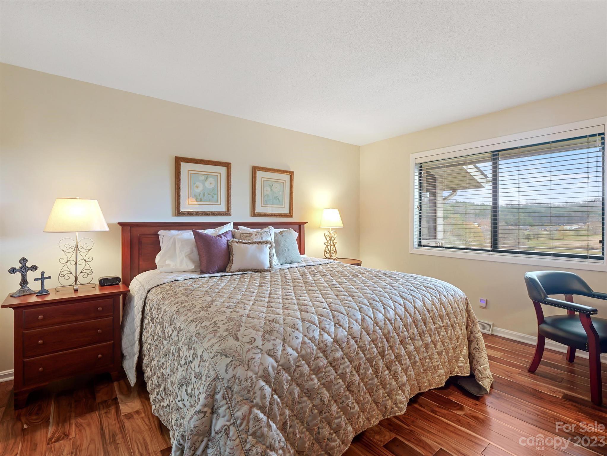 Furnished bedroom with a patterned quilt, hardwood floors, and a window overlooking homes and a wooded ridge.