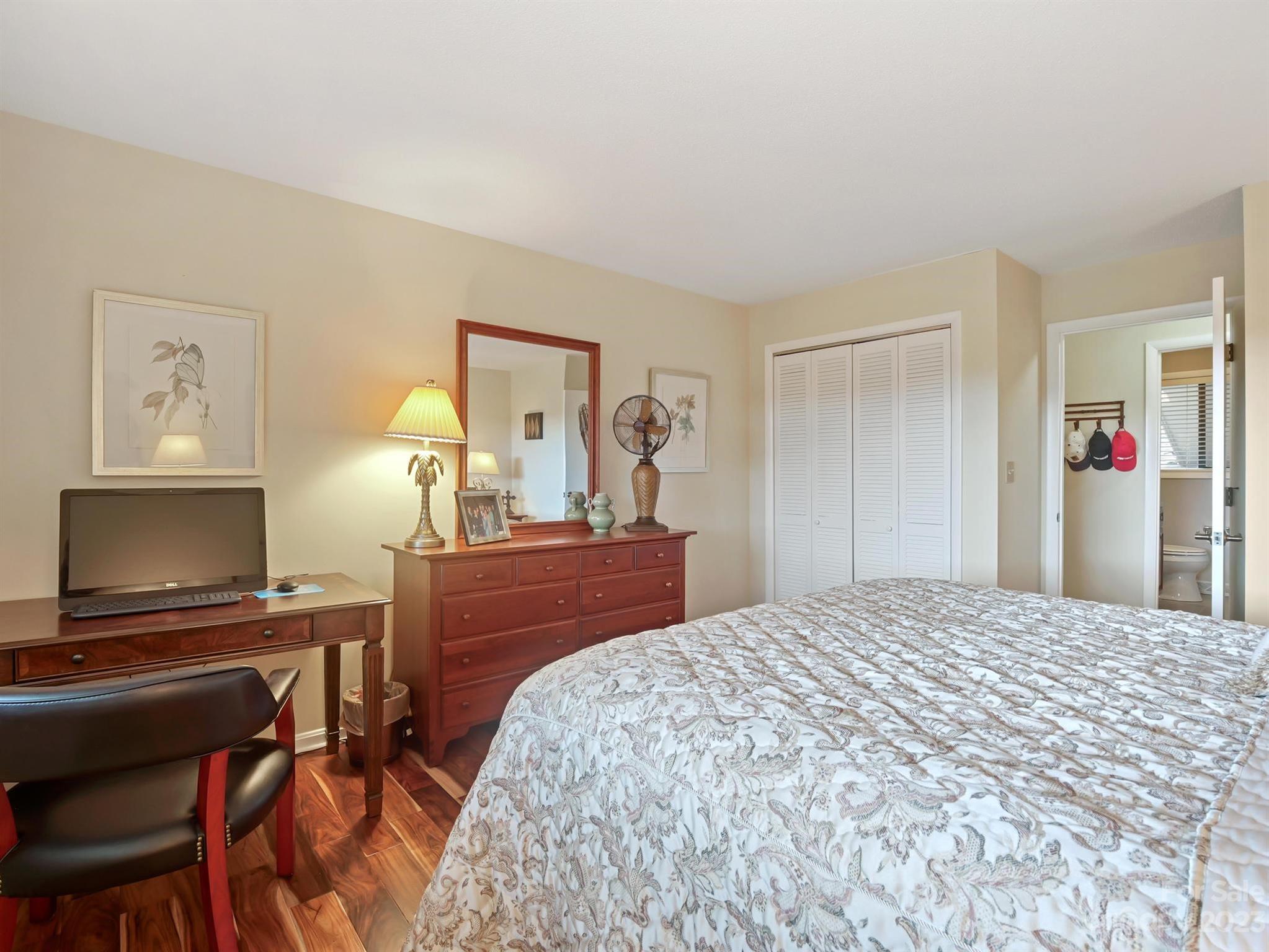 Furnished bedroom with a patterned bed, wooden dresser and mirror, and desk with computer.