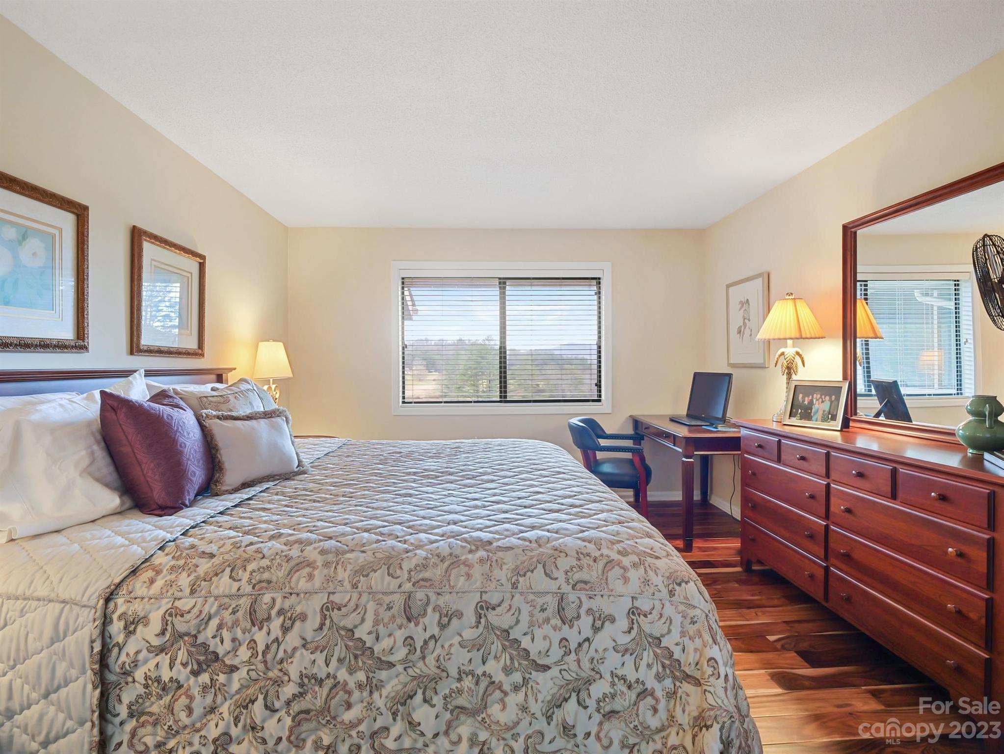 Furnished bedroom with a patterned bed, wood dresser, desk, and hardwood floors.
