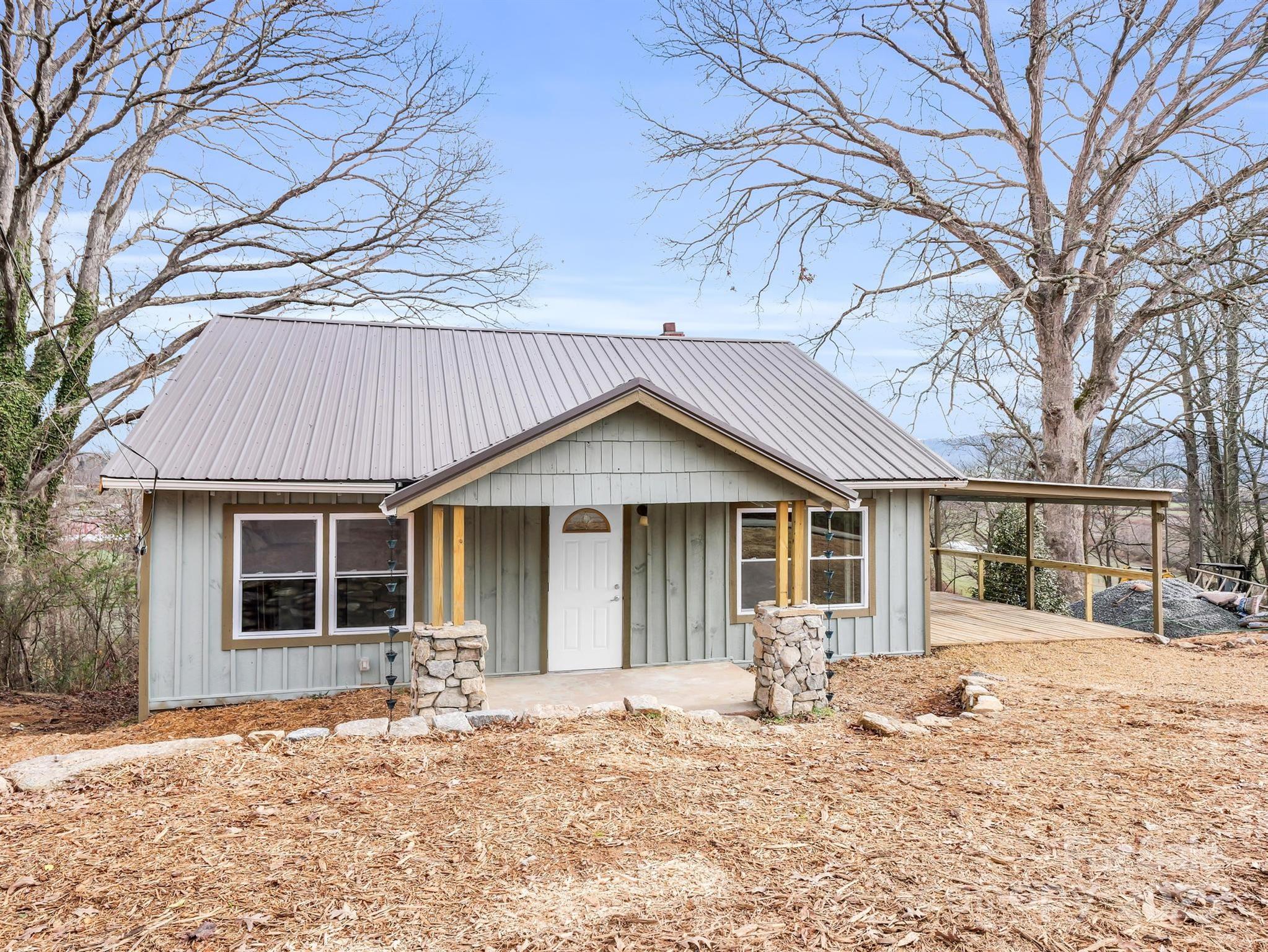 Light-gray house with a metal roof, stone porch pillars, and a side deck beneath leafless trees.
