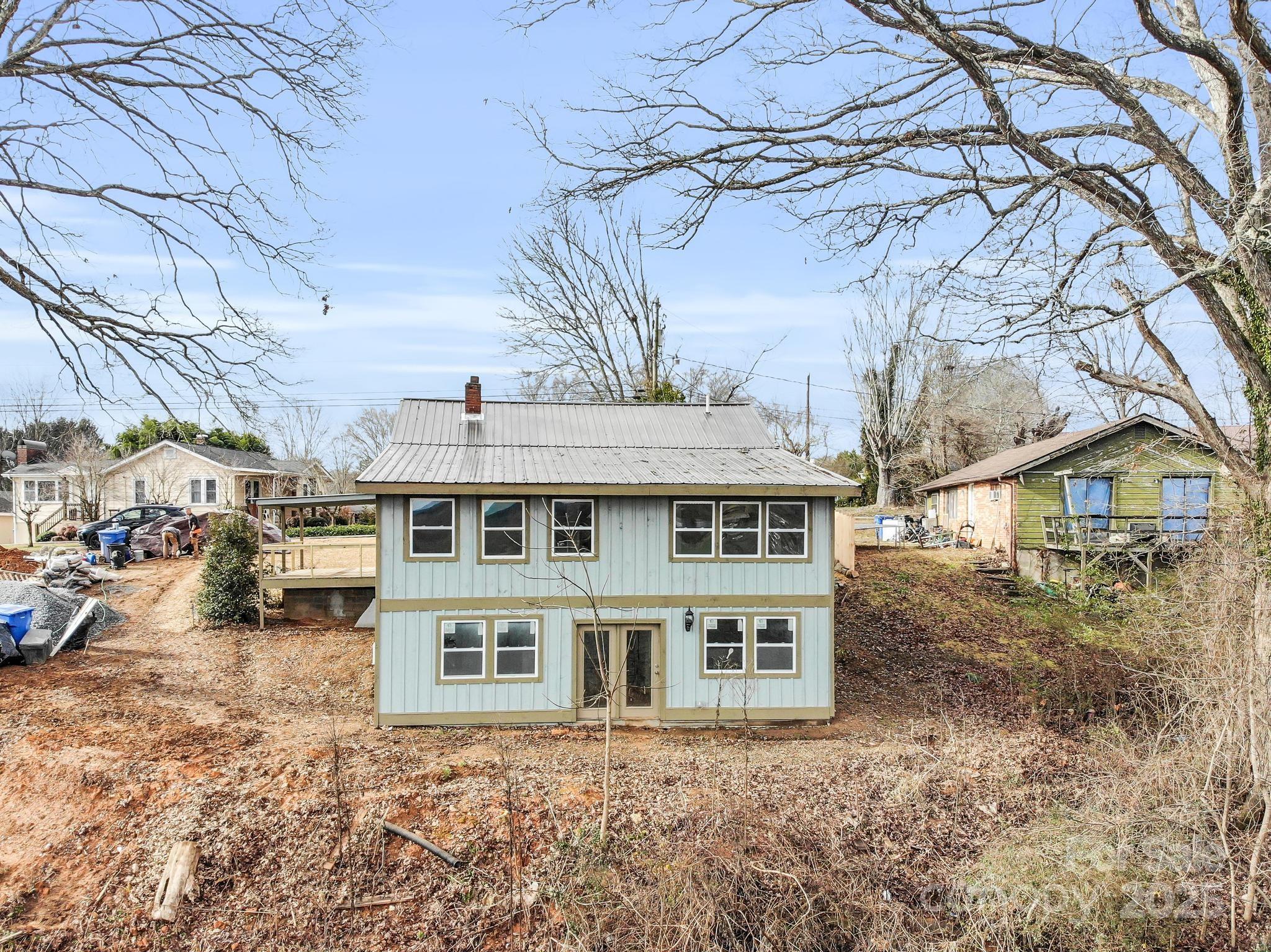 Two-story pale blue-gray house with a metal roof and wooden side deck, framed by leafless trees.