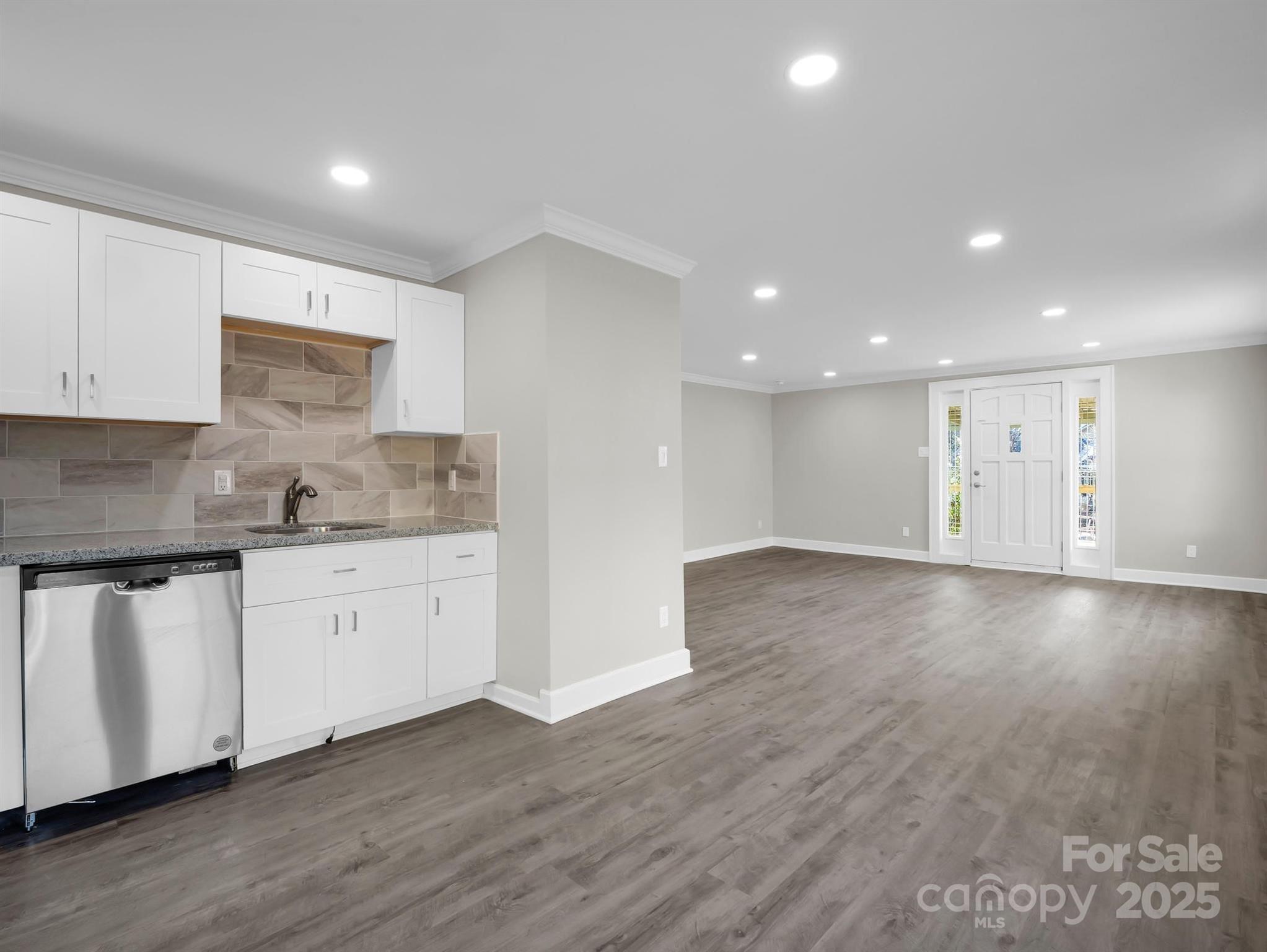 Open kitchen and living area with white cabinetry, gray tile backsplash, and wood-look floors.
