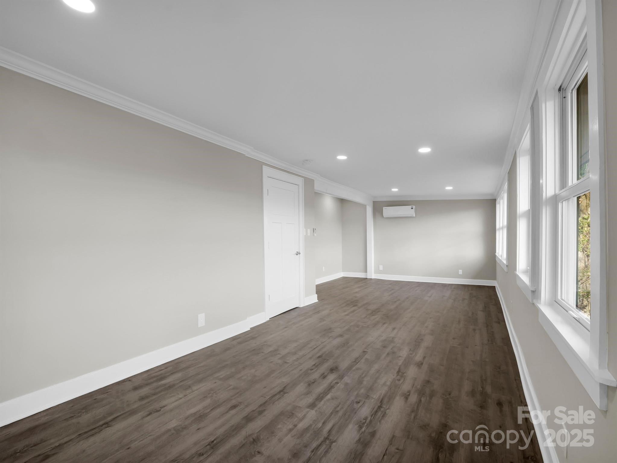 Empty room with dark wood-look floors, recessed lights, and large windows along one wall.