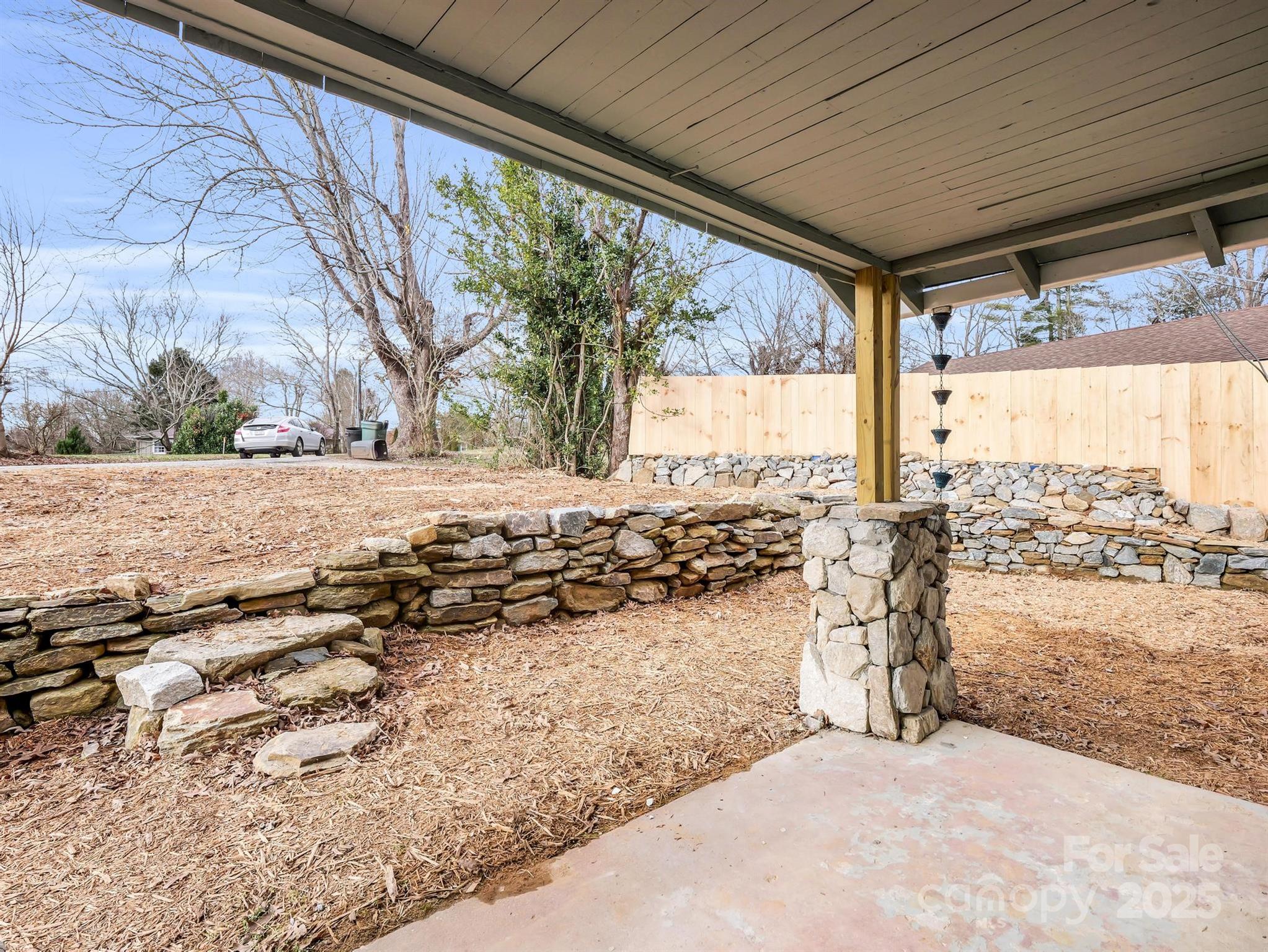 Covered backyard patio with a stone column, tiered stone walls, and a tall wooden fence.