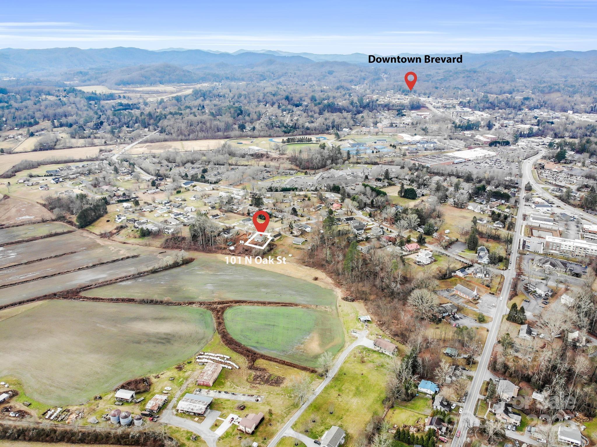 Aerial view of 101 N Oak Street, with its lot outlined amid open fields and homes, and mountains beyond Brevard.