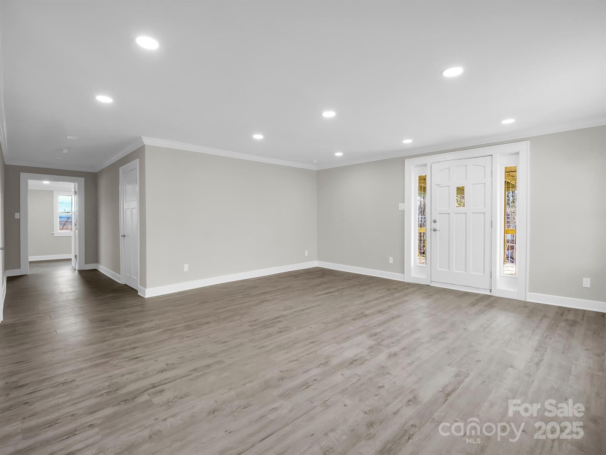 Empty room with gray wood-look floors, recessed lights, and a white front door with sidelights.