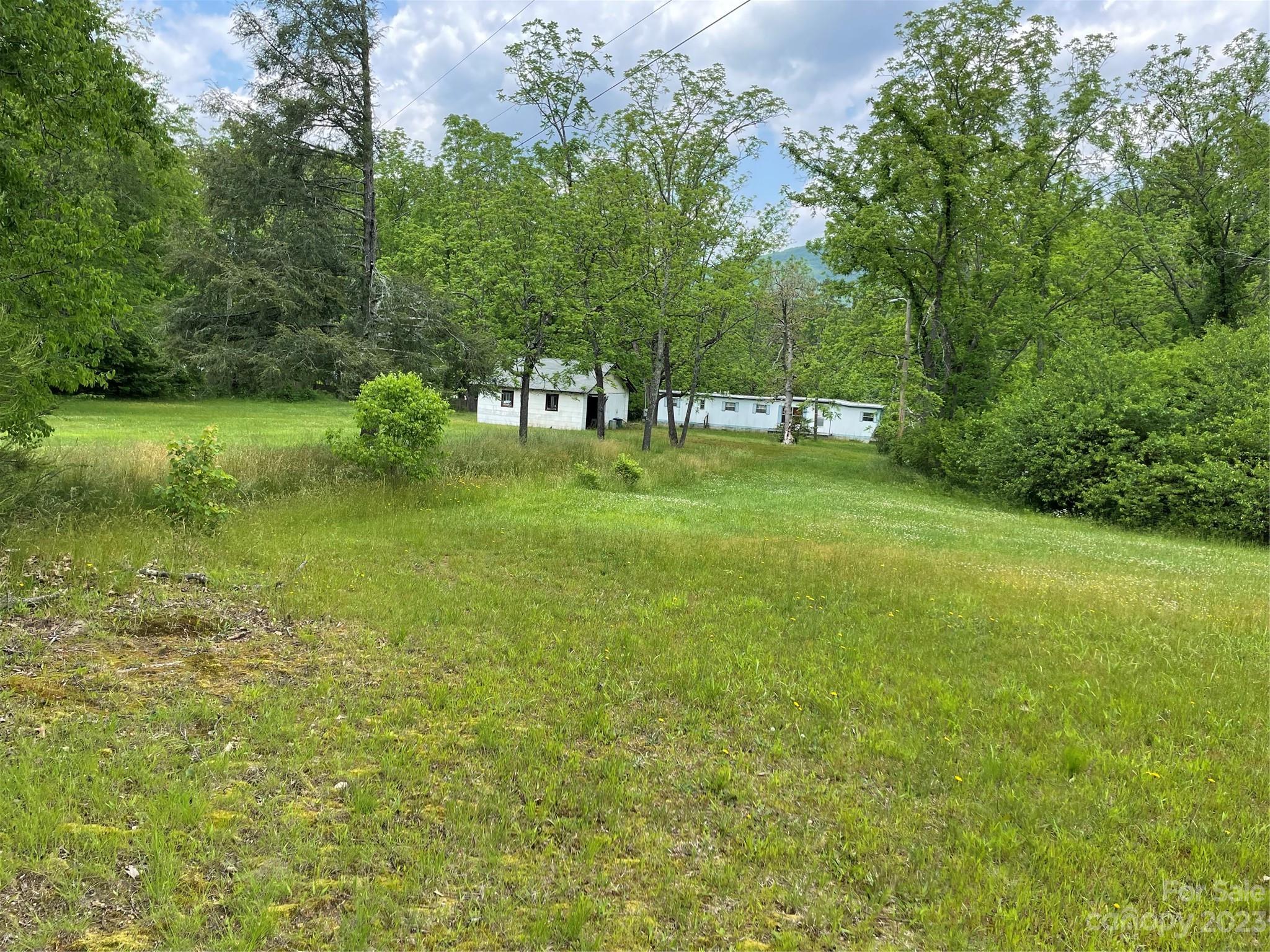 White manufactured homes sit beyond a broad grassy yard, surrounded by mature trees.