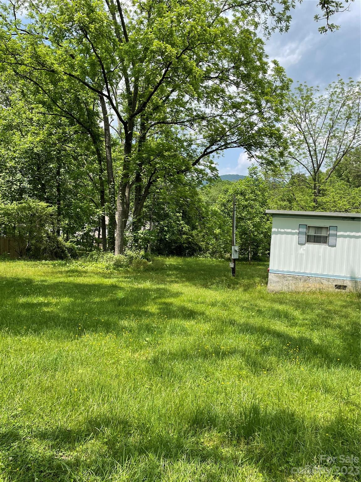 Grassy yard shaded by mature trees, with part of a manufactured home and a distant mountain ridge.