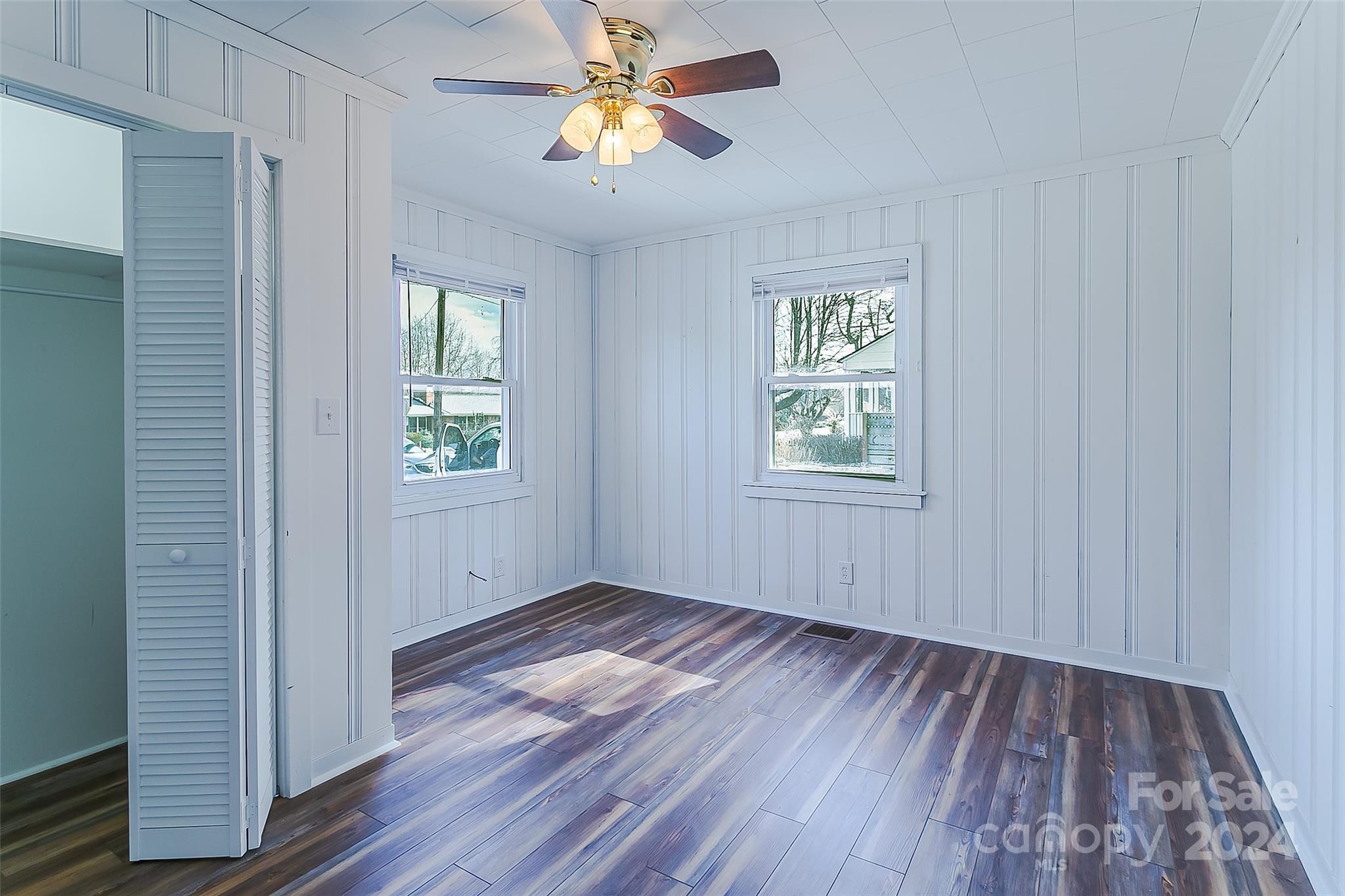 Empty room with dark plank flooring, white paneled walls, two windows, and an open louvered closet door.
