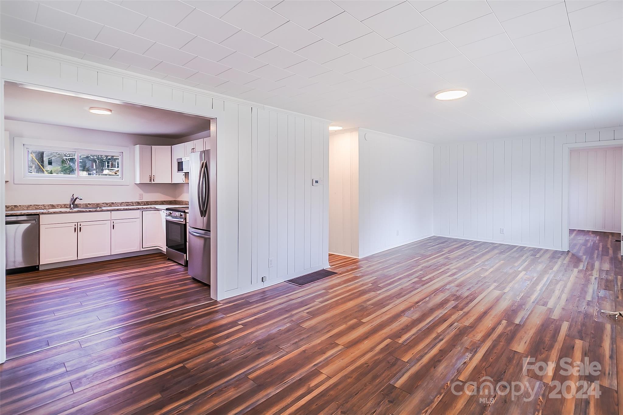 Vacant living area with multicolored wood-look flooring and an adjoining white kitchen.