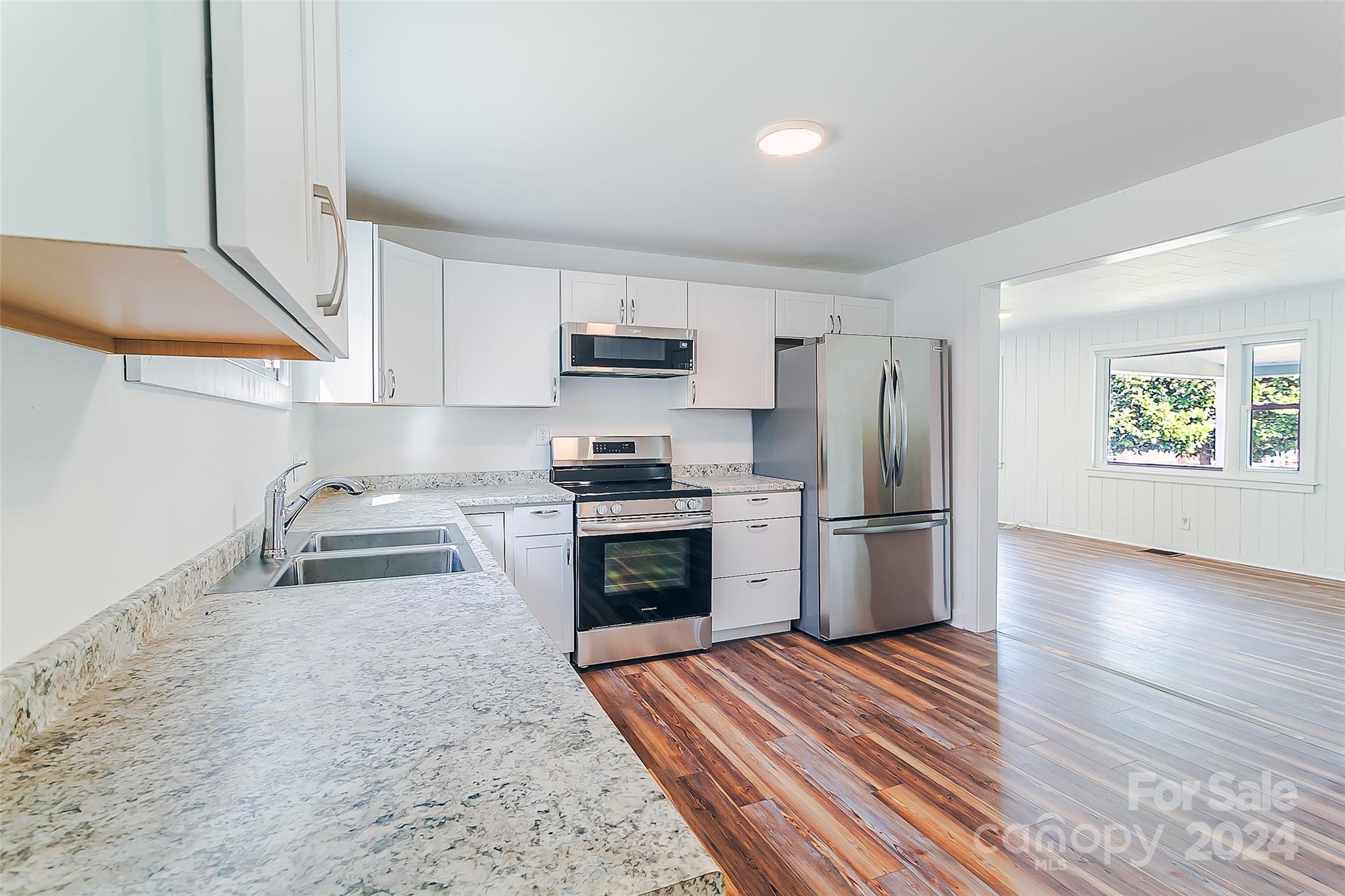 Vacant kitchen with granite counters, stainless steel appliances, and wood-look floors opening to a bright adjoining room.