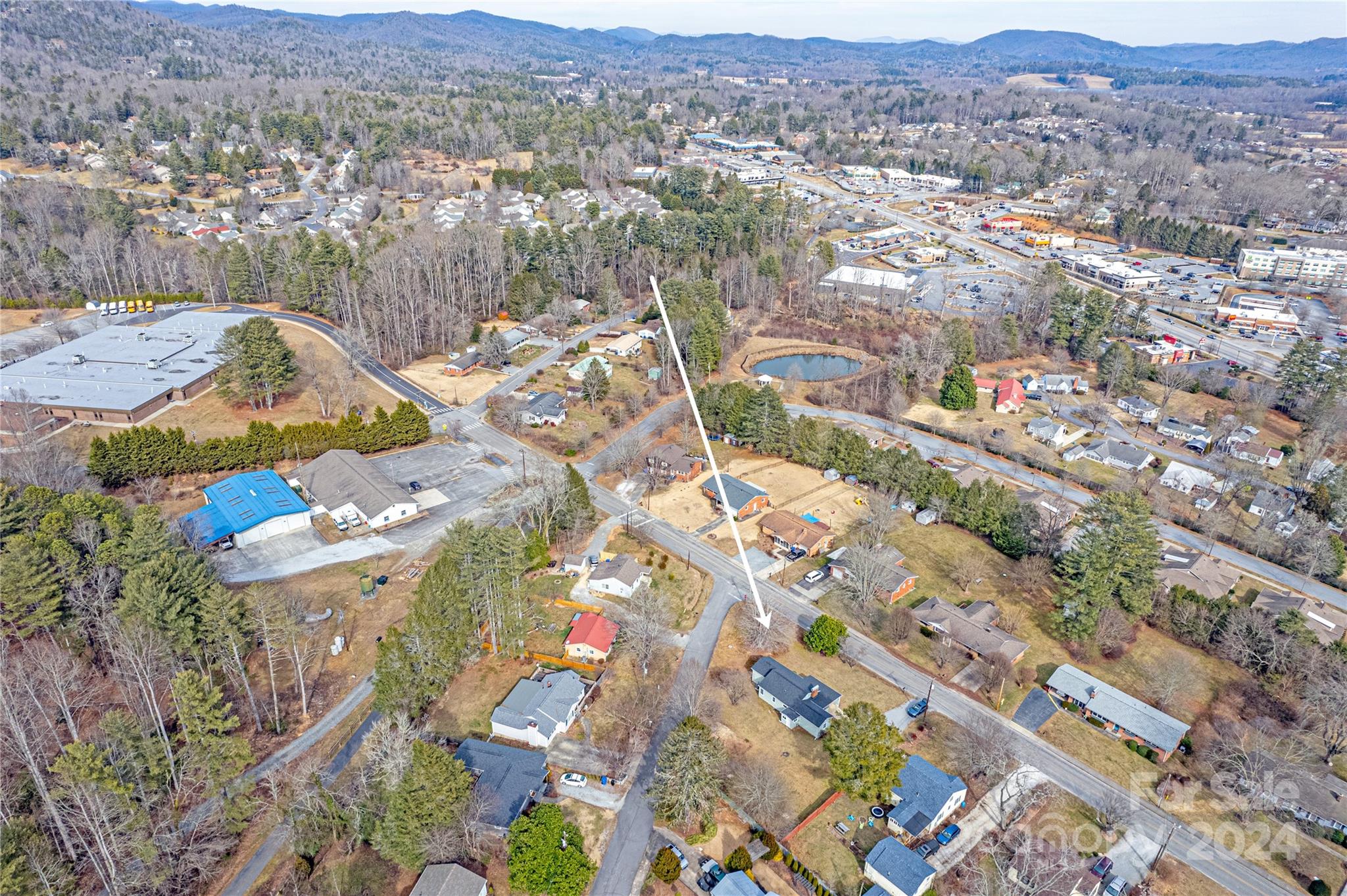 Aerial neighborhood view with a white arrow pointing to the property among homes and wooded mountains.