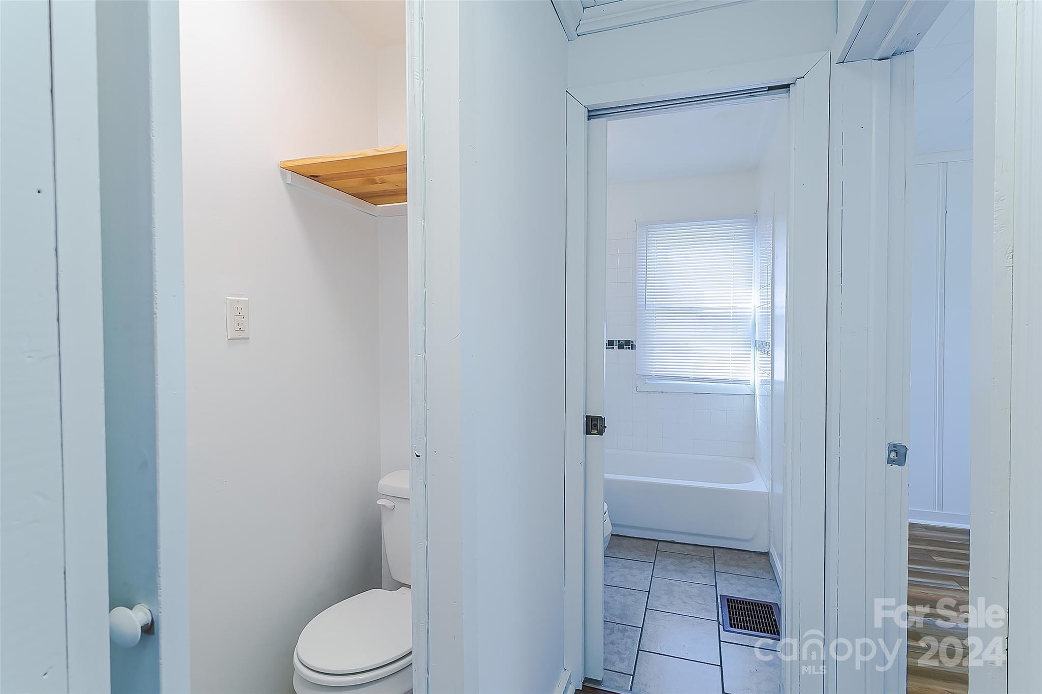 Bathroom with a white bathtub beneath a blinded window and gray tile flooring.