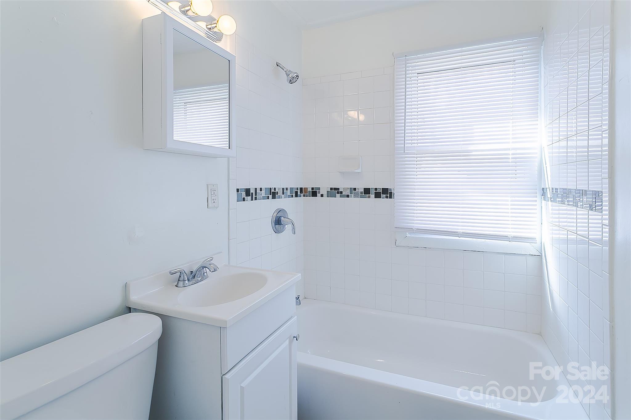 Bathroom with white bathtub and tiled shower, mosaic accent strip, window blinds, vanity, and toilet.