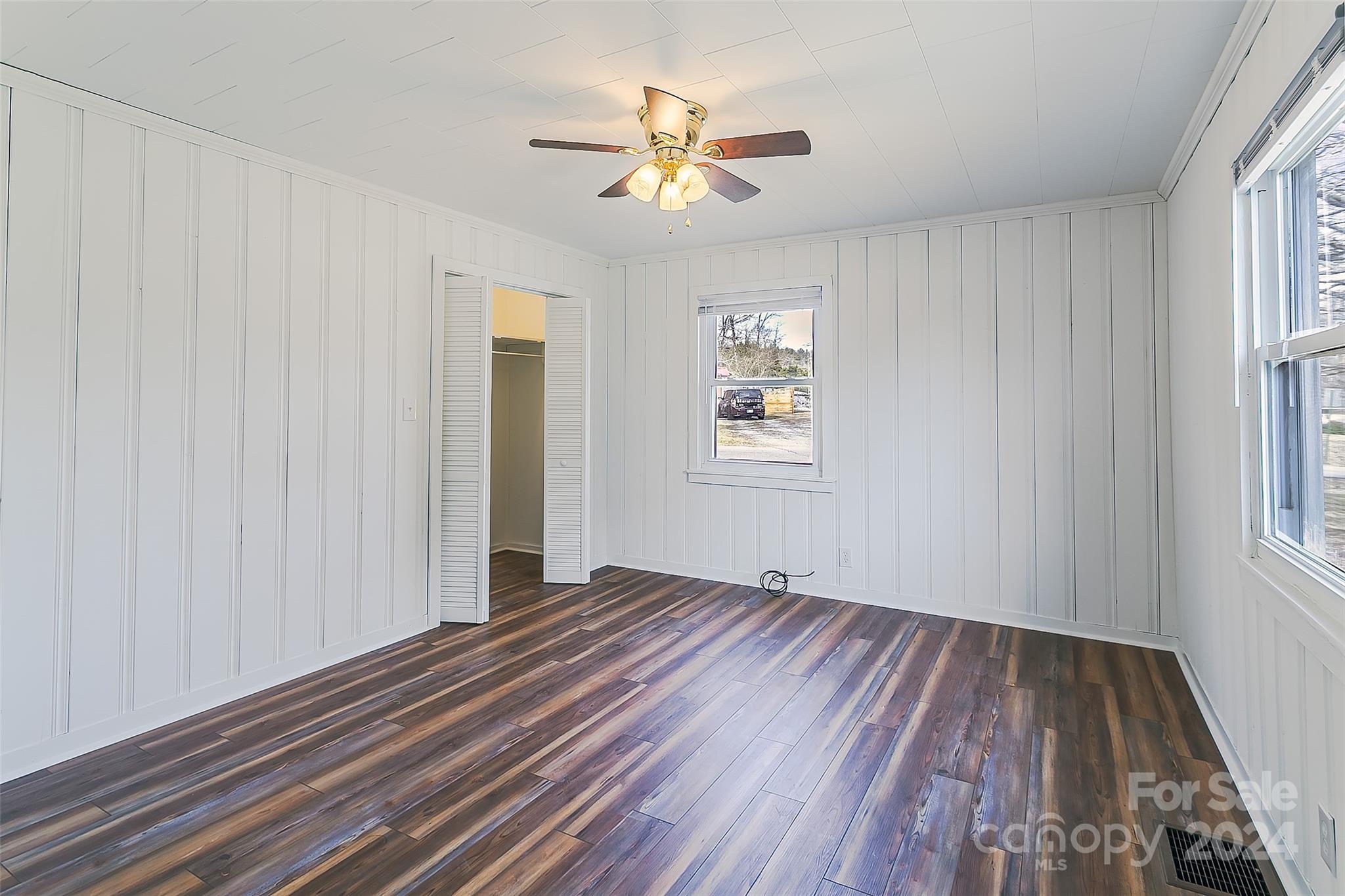 Vacant bedroom with white paneled walls, dark plank flooring, ceiling fan, and an open closet.