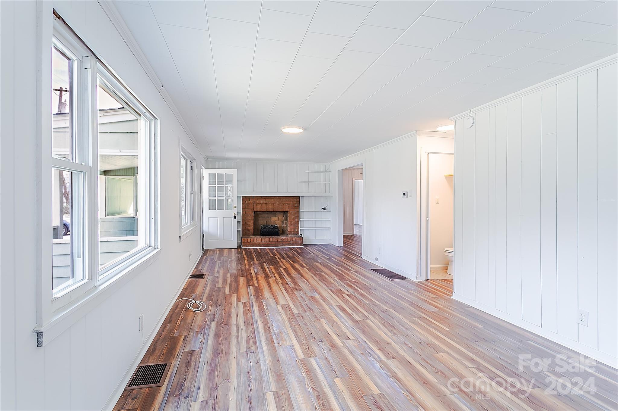 Empty living room with wide windows, wood-look flooring, built-in shelves, and a brick fireplace.