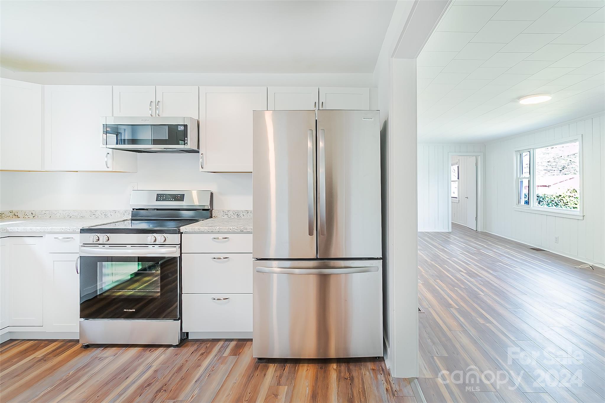 Bright white kitchen with stainless steel appliances and wood-look floors opening to an empty paneled room.