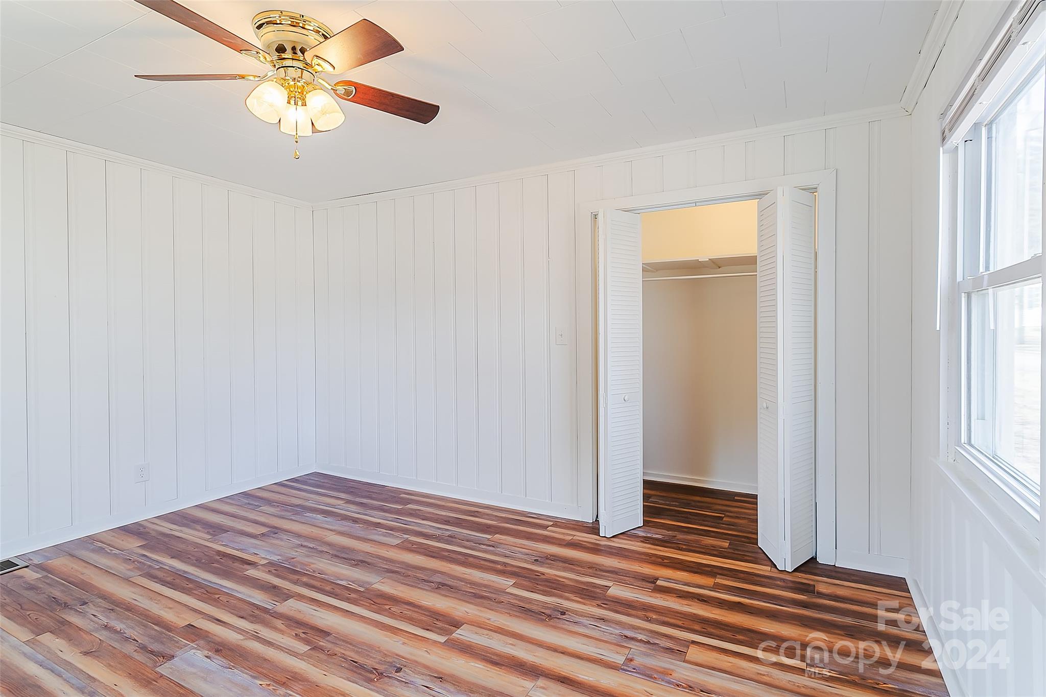 Empty bedroom with wood-look flooring, a ceiling fan, large windows, and open closet doors.