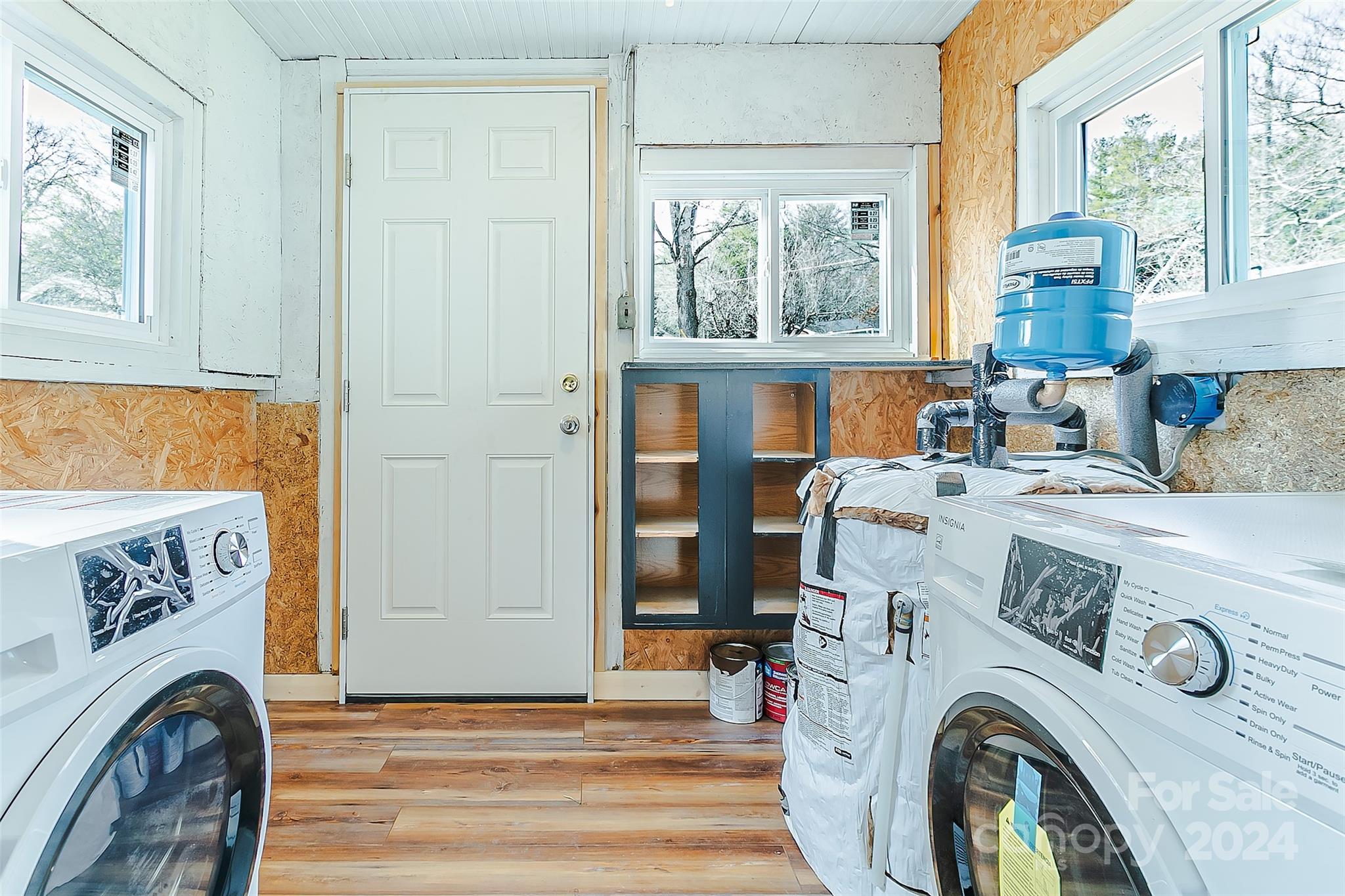Laundry room with front-loading washer and dryer, a blue water filtration tank, and unfinished OSB walls.