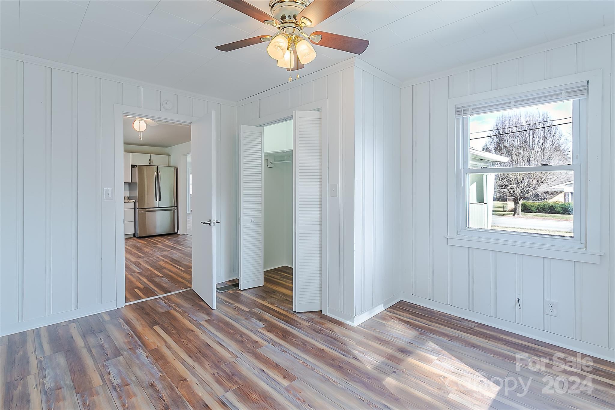 Vacant bedroom with wood-look flooring, white paneled walls, and a window overlooking a leafless tree.