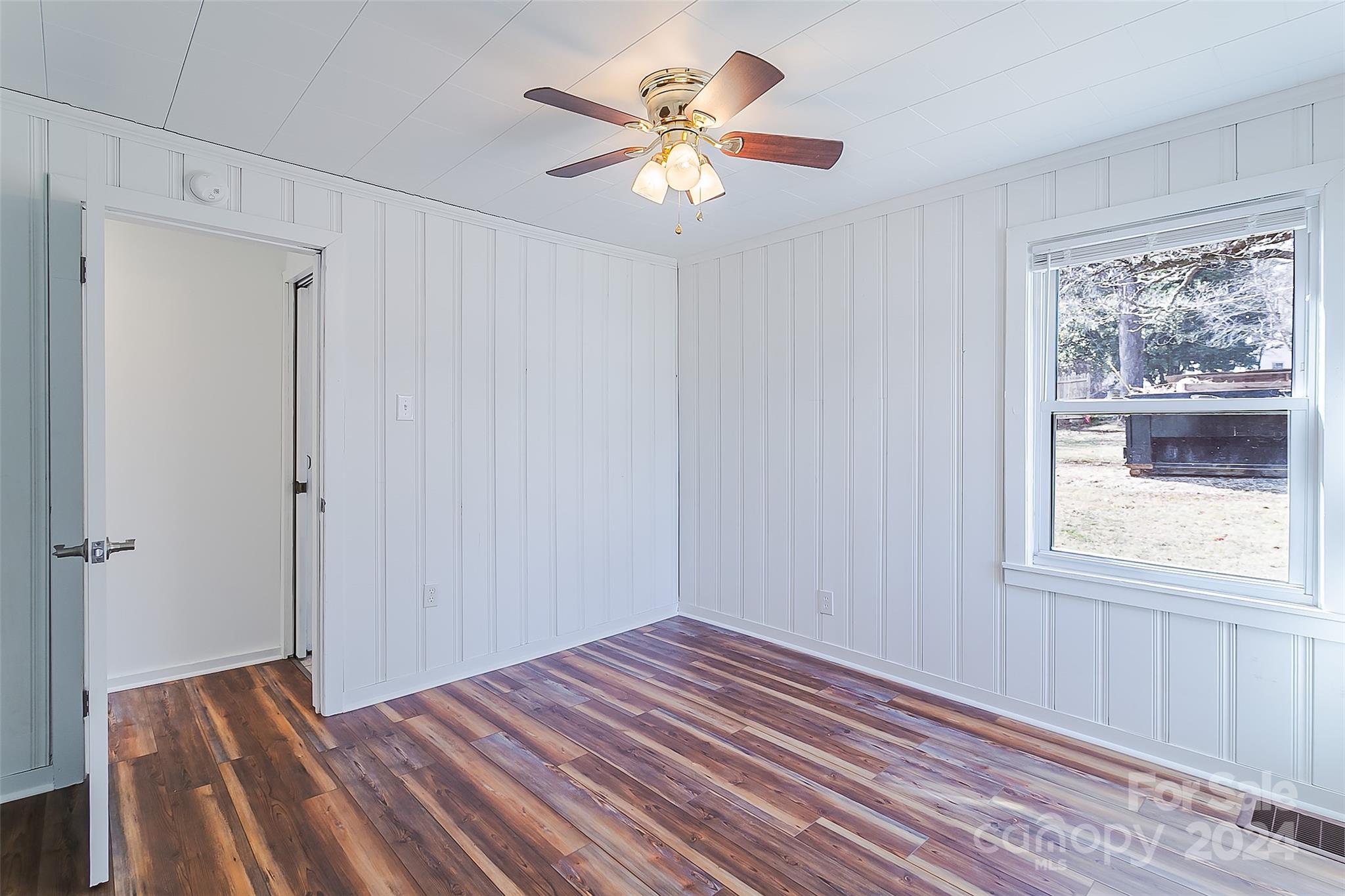 Empty room with white paneled walls, wood-look flooring, a ceiling fan, and a large window.