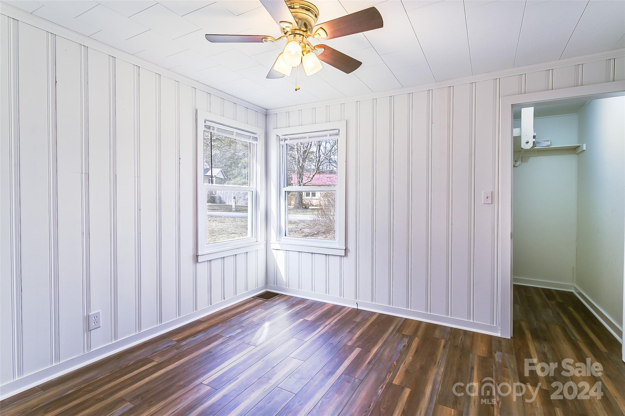 Vacant bedroom with two corner windows, paneled walls, dark wood-look floors, and a ceiling fan.