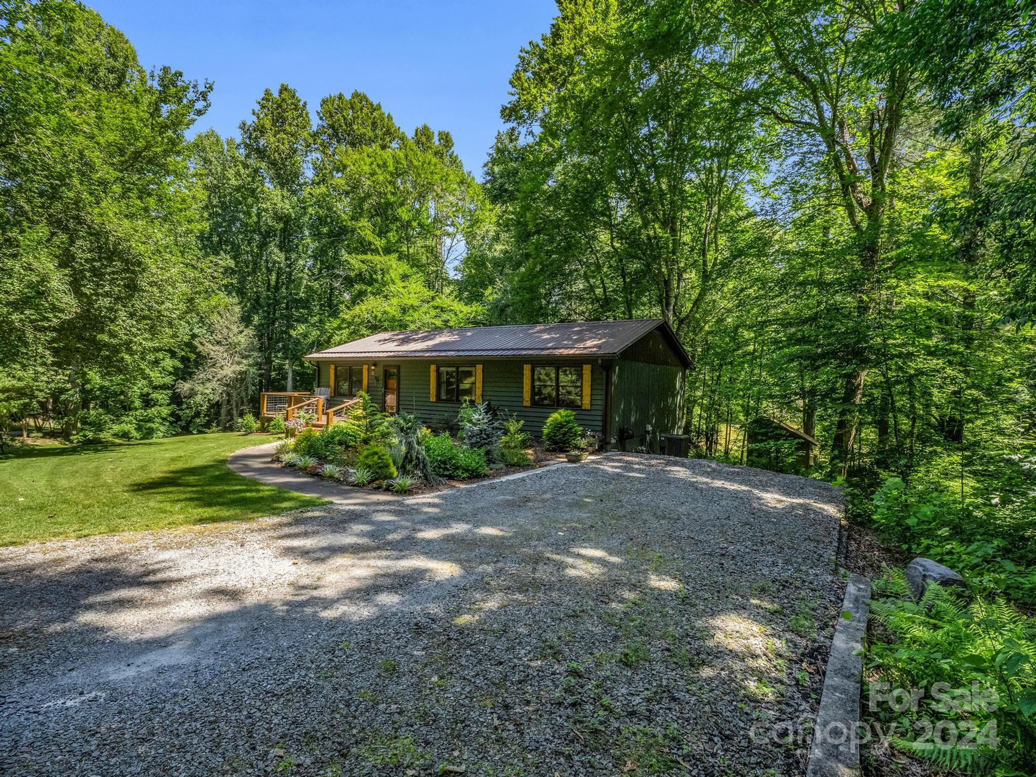 Dark green single-story home with yellow shutters, a wooden deck, and a gravel driveway in a wooded setting.