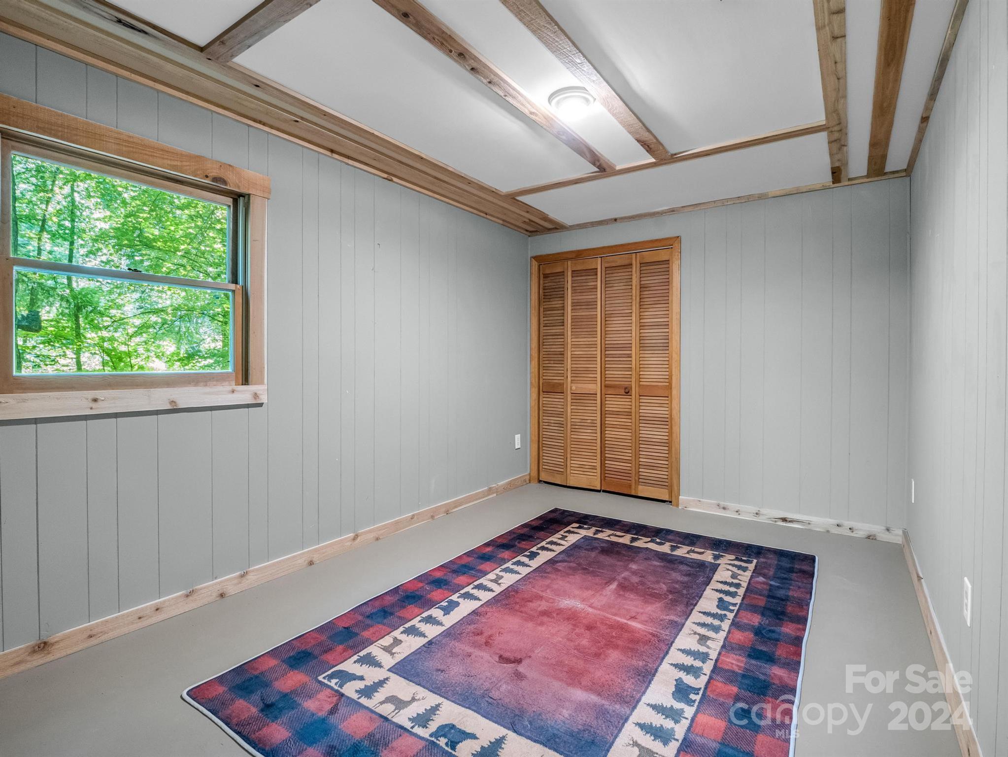 Empty room with exposed wood ceiling beams, louvered closet doors, and a window overlooking trees.