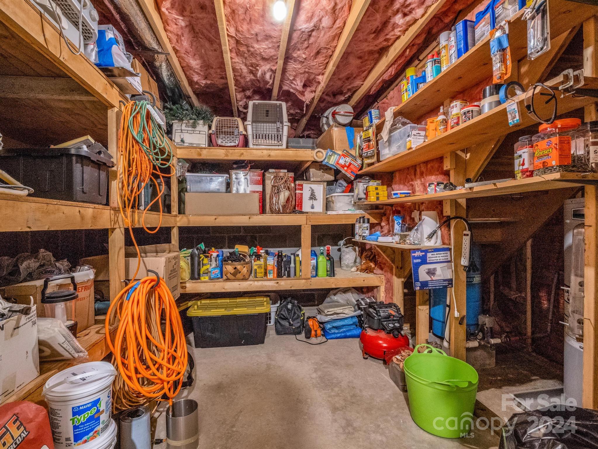 Unfinished storage room with exposed insulation, packed wooden shelves, orange extension cords, and tools.