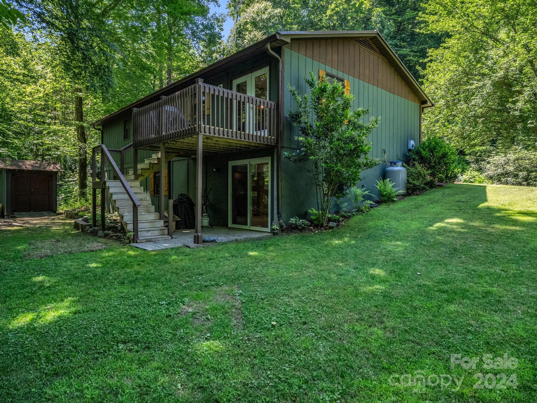 Green house with a raised deck and exterior stairs, set beside a broad lawn and wooded surroundings.