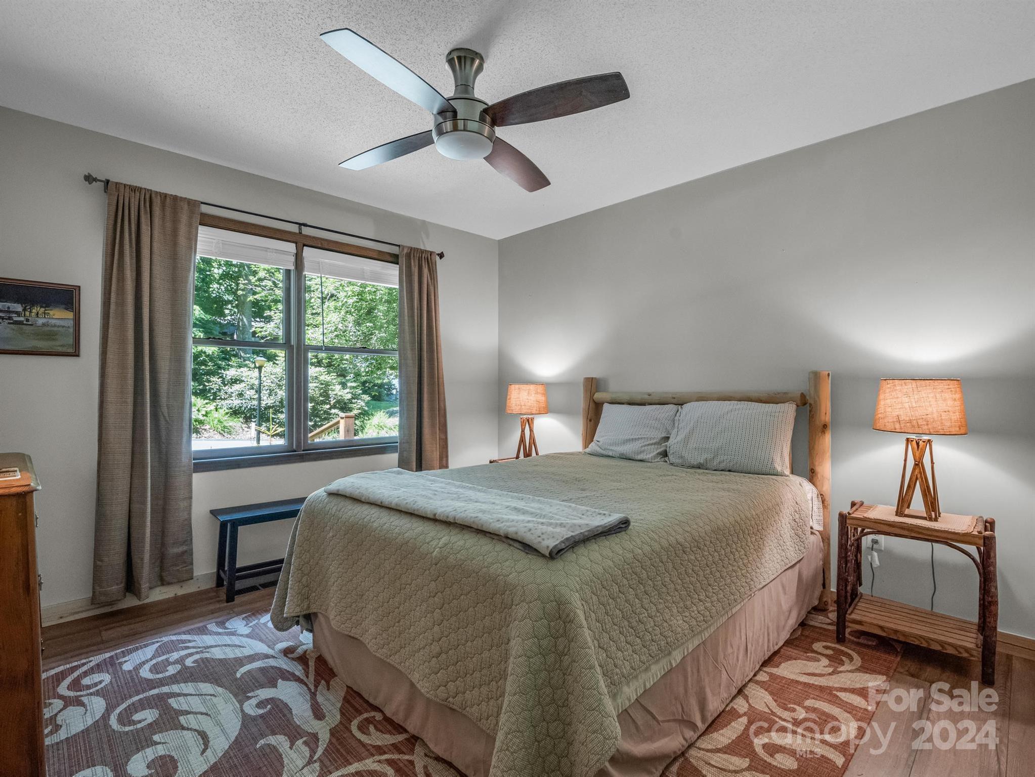Furnished bedroom with a large bed, wooden headboard, and wide window overlooking leafy greenery.