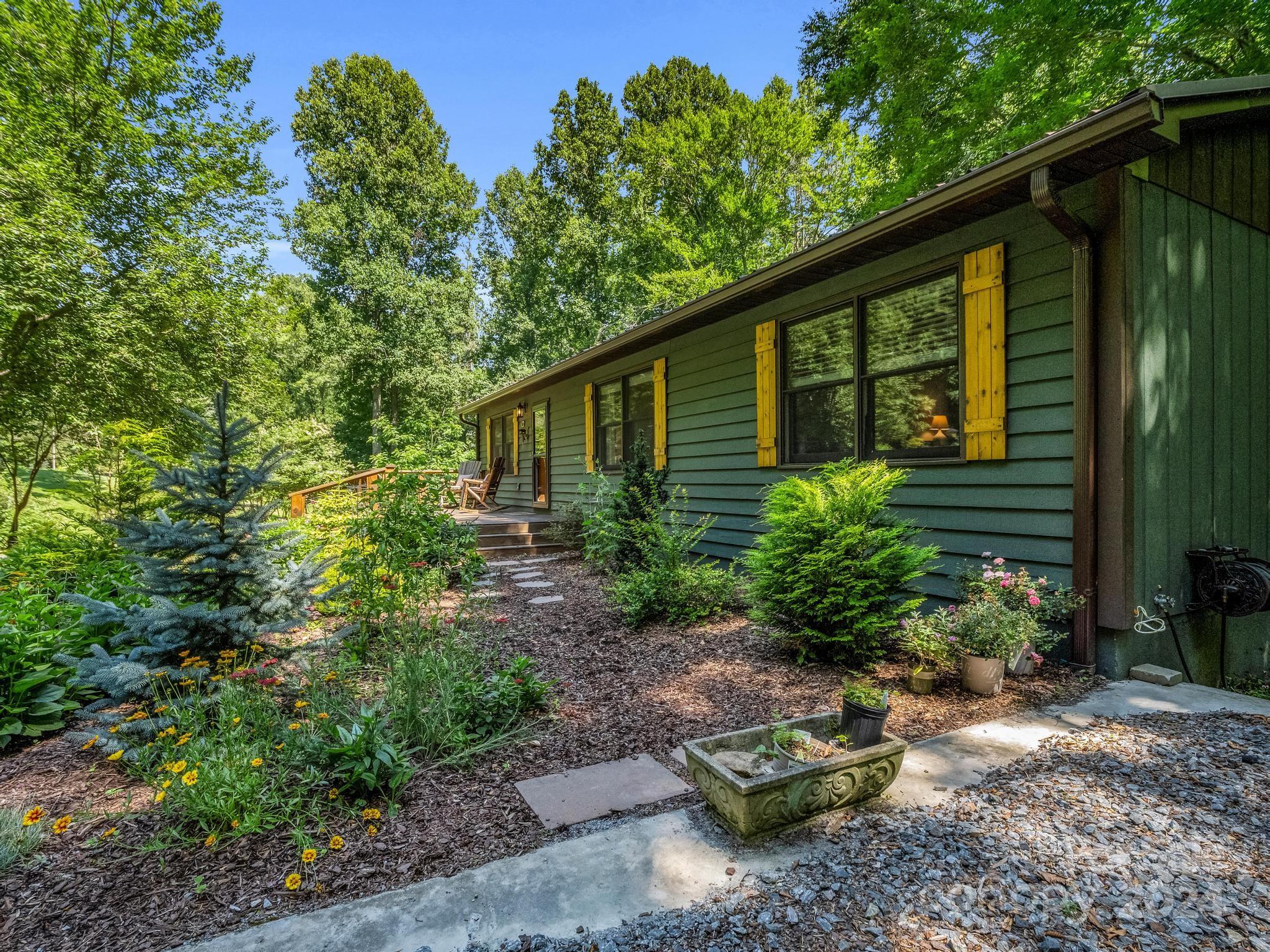 Green home with yellow shutters beside a garden path and small deck surrounded by trees.
