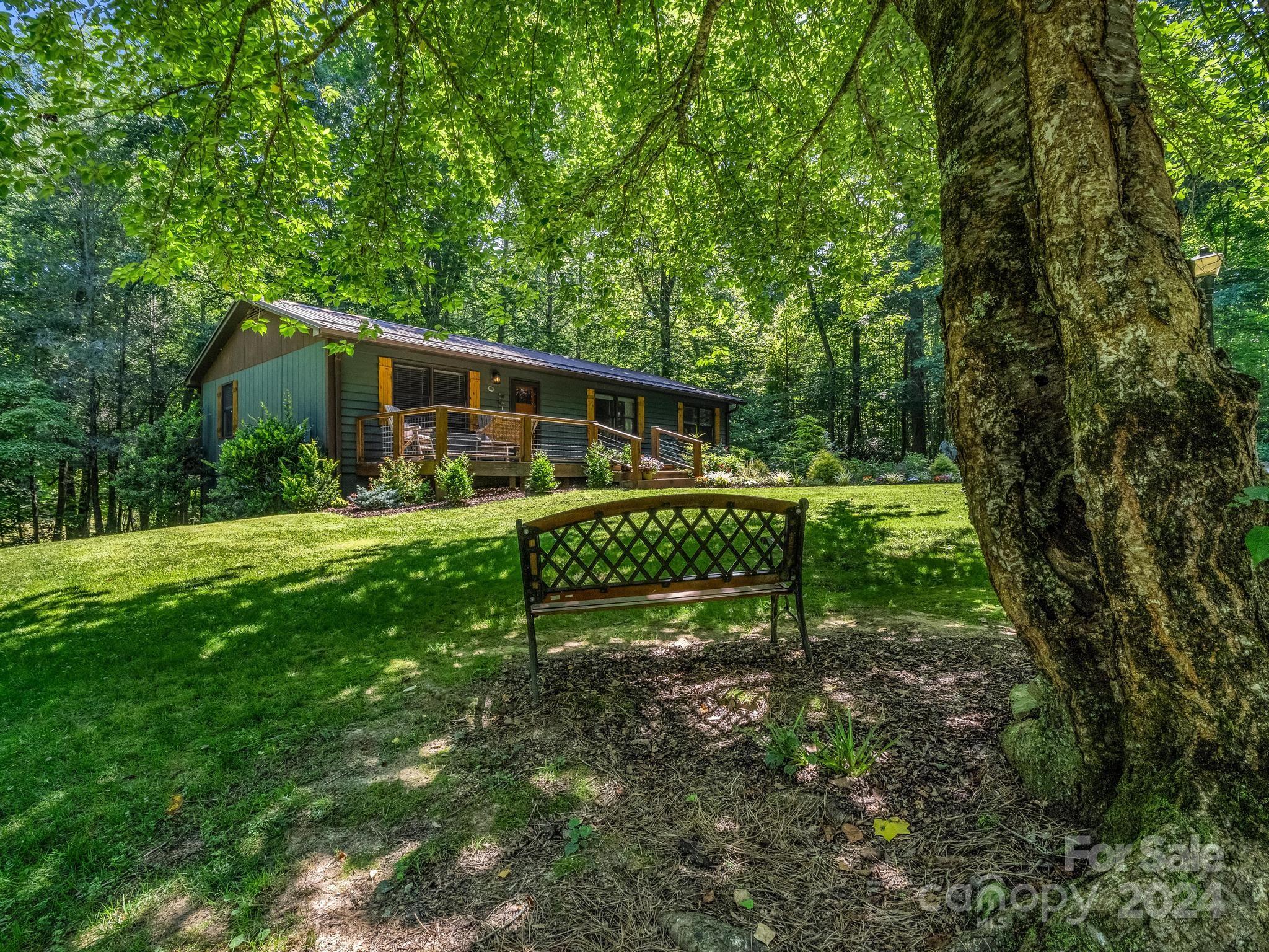 Green home with a raised deck in a wooded clearing, seen beyond a lawn bench.
