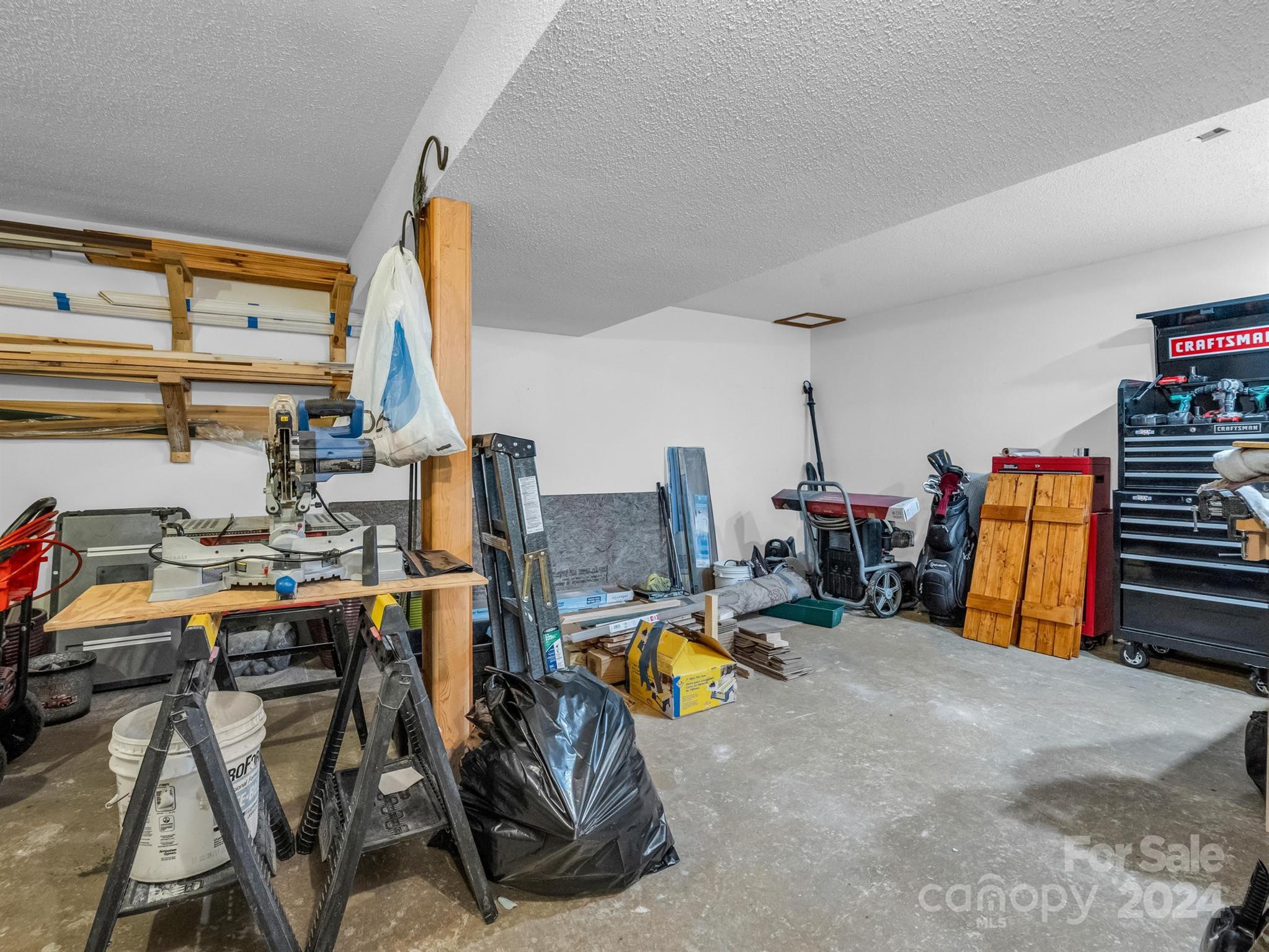 Garage workshop with a miter saw, stored lumber, ladders, tool chests, and golf bags.