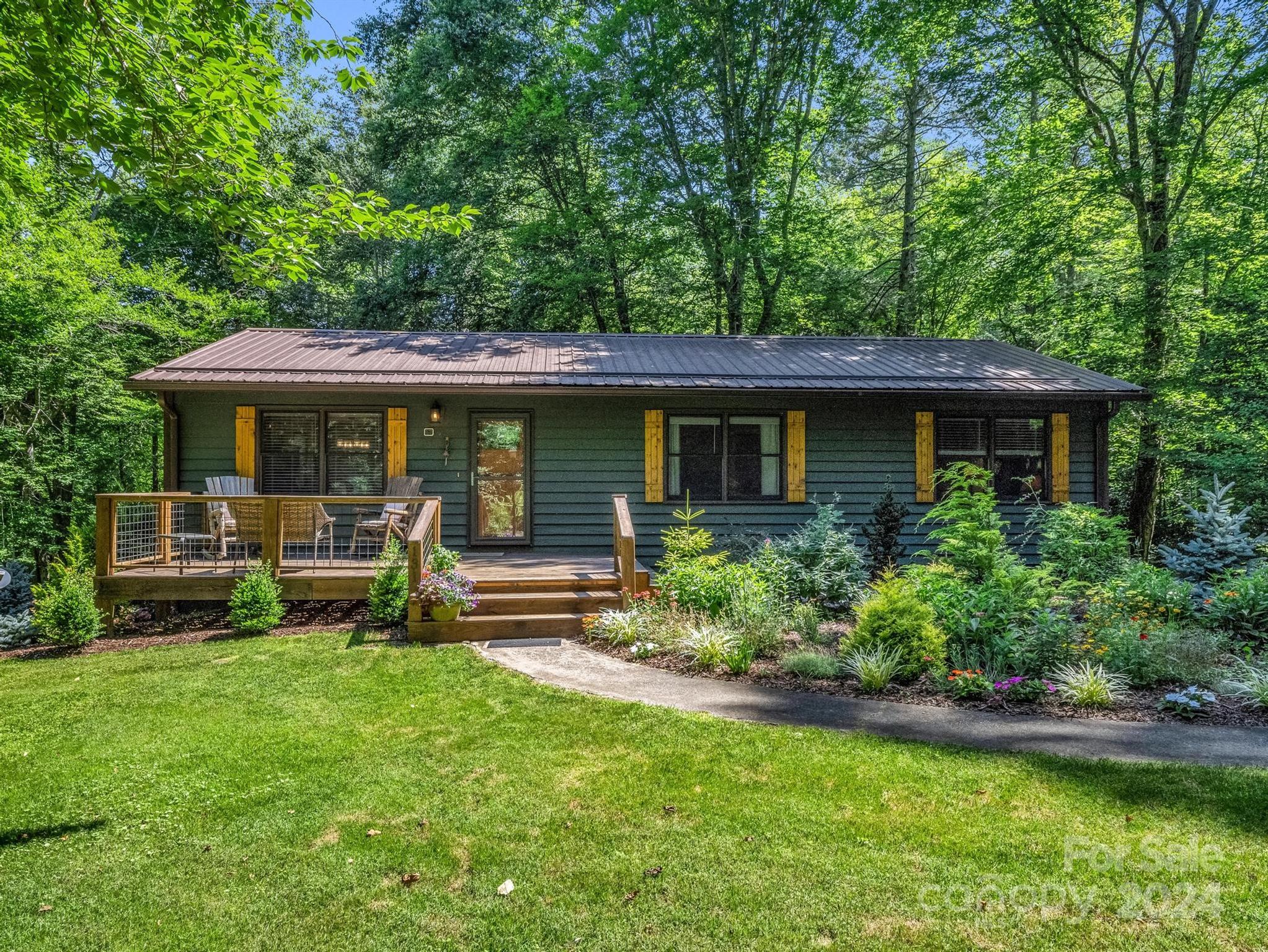 Dark green home with yellow shutters and a raised deck surrounded by lush trees and garden beds.