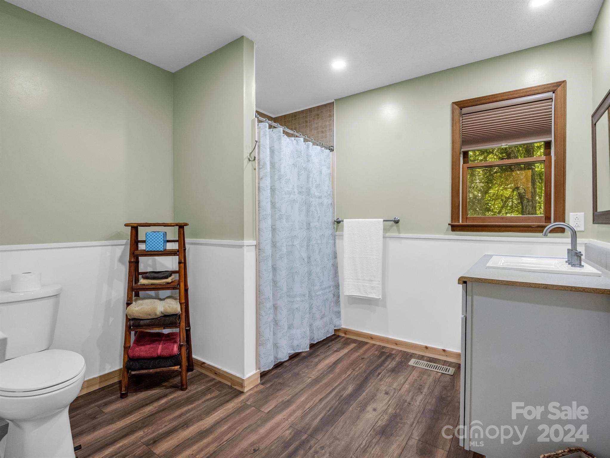 Bathroom with a shower curtain, wood-look flooring, and a wood-trimmed window overlooking trees.