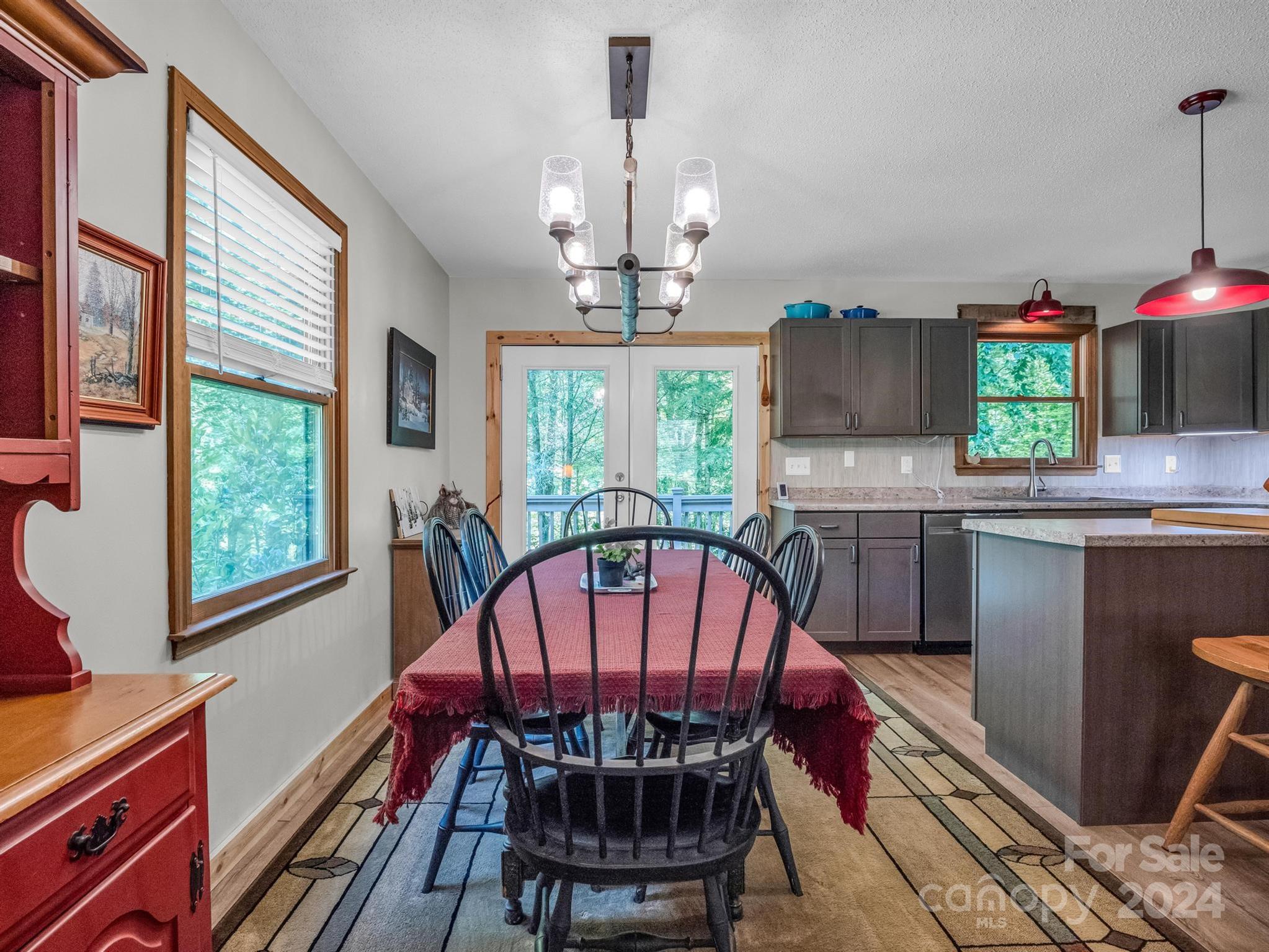 Dining area with red tablecloth opens to a gray-cabinet kitchen and French doors overlooking greenery.
