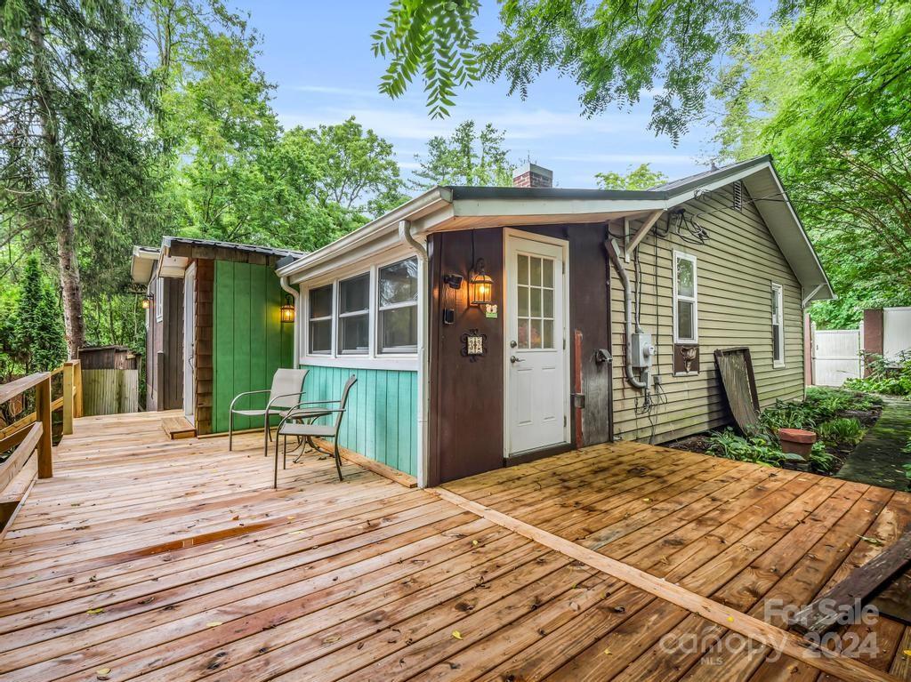 Small house with a broad wooden deck and chairs, surrounded by leafy trees.