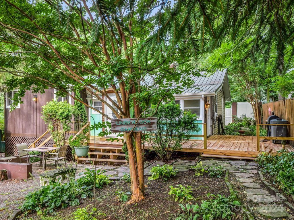 Wooded backyard with a raised deck, stone path, patio furniture, and a garden sign.