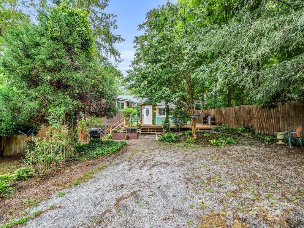 Fenced gravel yard leading to a small house with a raised deck beneath mature trees.