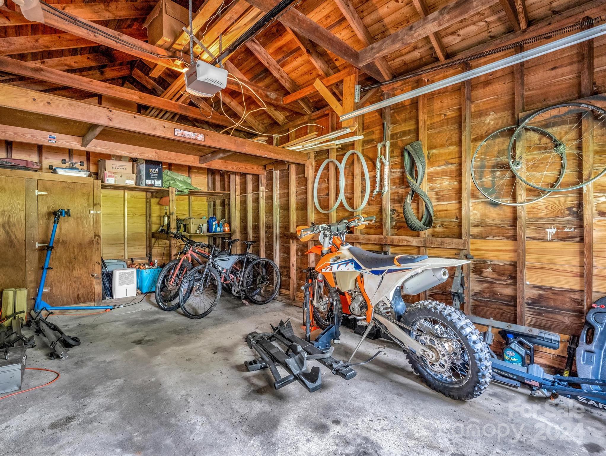 Wood-framed garage with exposed rafters, several bicycles, and an orange-and-white dirt bike.