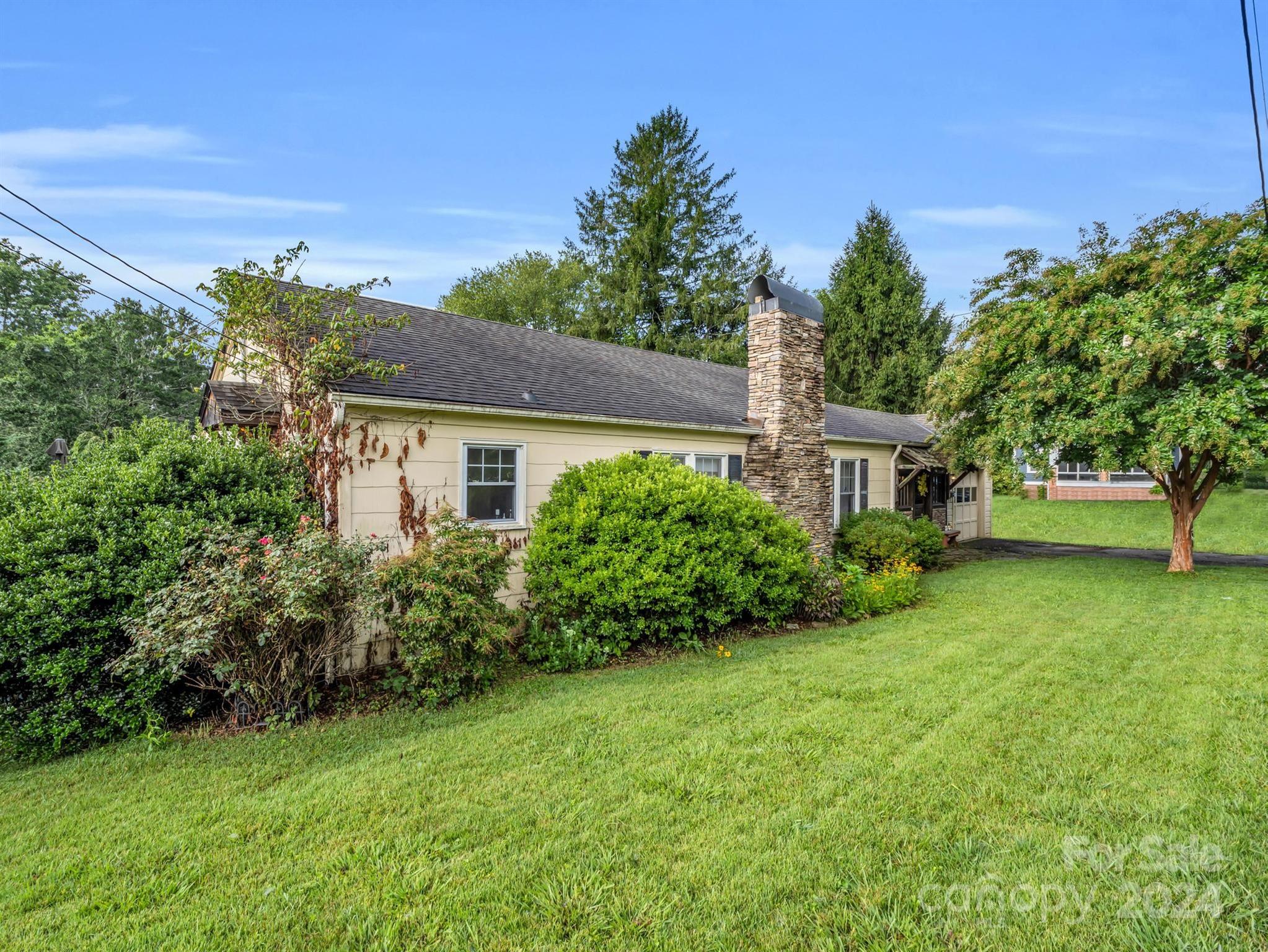 Single-story cream home with a tall stone chimney, landscaped shrubs, and a broad lawn.