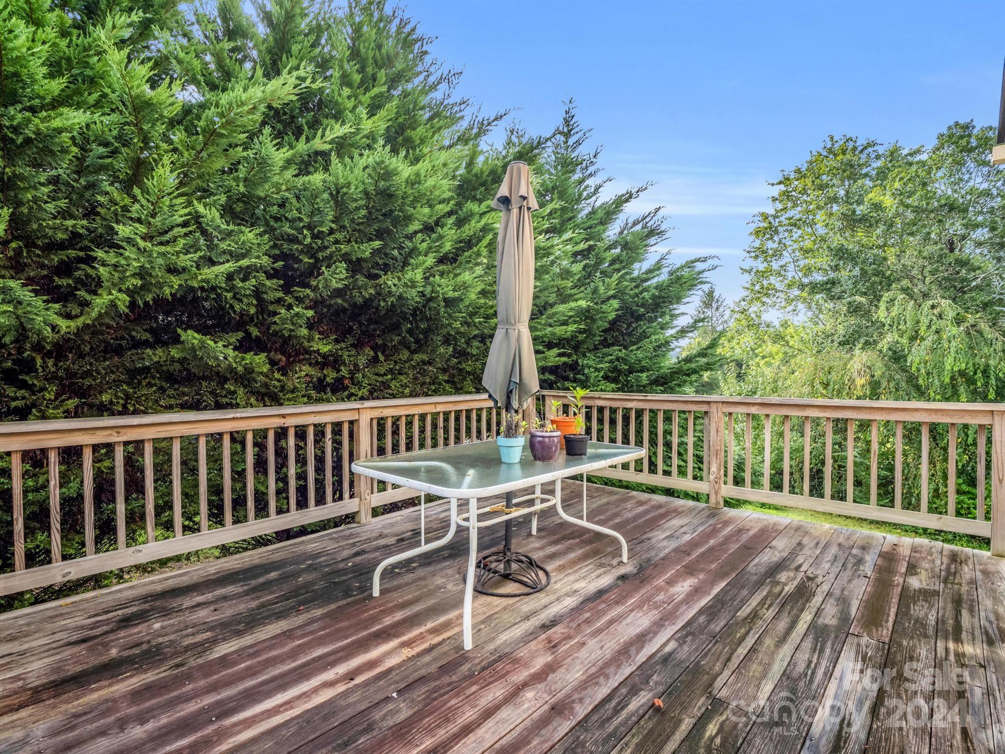 Wooden deck with a glass-top table, potted plants, and closed umbrella overlooking dense greenery.