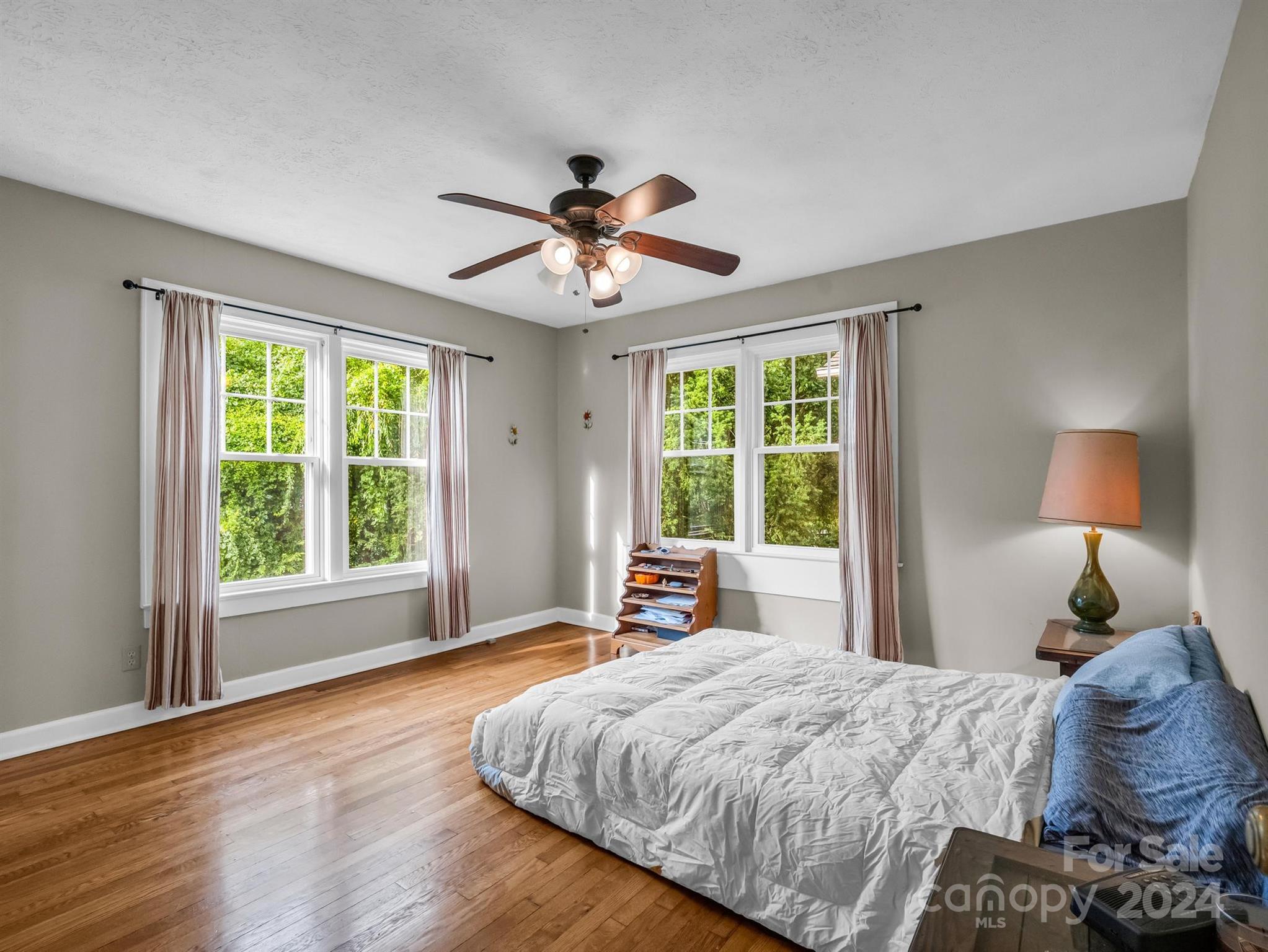Bedroom with hardwood floors, a ceiling fan, and large windows overlooking greenery.