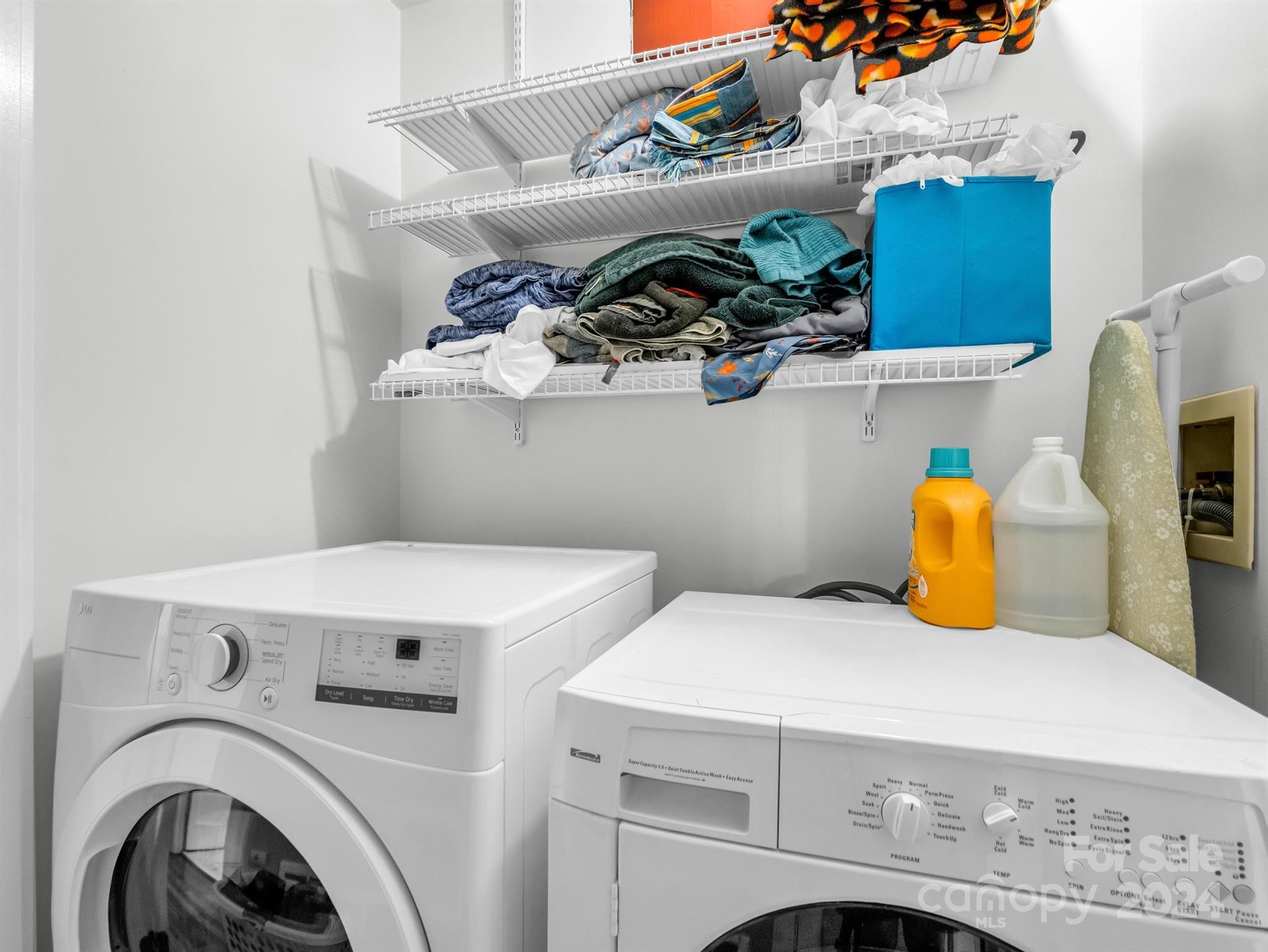 Laundry room with front-loading washer and dryer beneath wire shelves stocked with laundry.