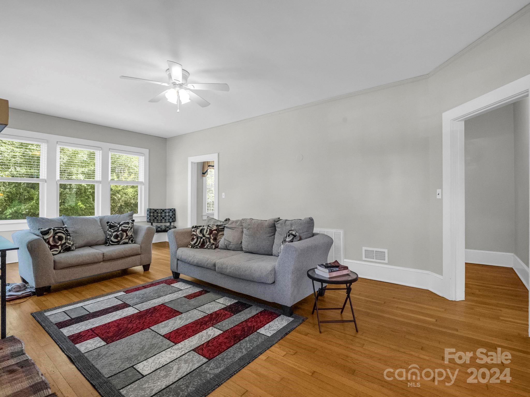 Living room with two gray sofas, a red-and-gray patterned rug, hardwood floors, and windows facing greenery.