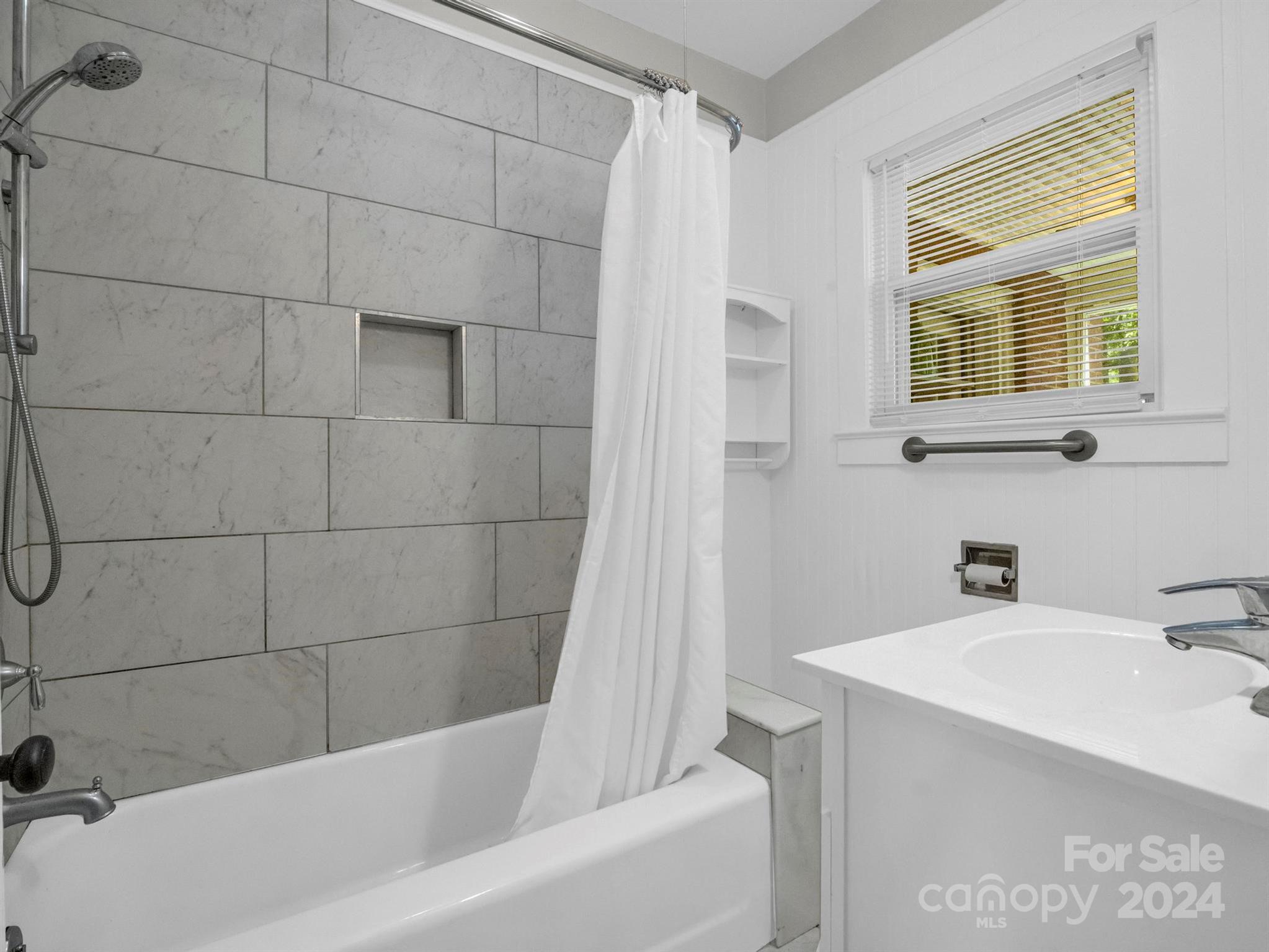 Bathroom with a tiled tub and shower, white curtain, vanity, and window with blinds.