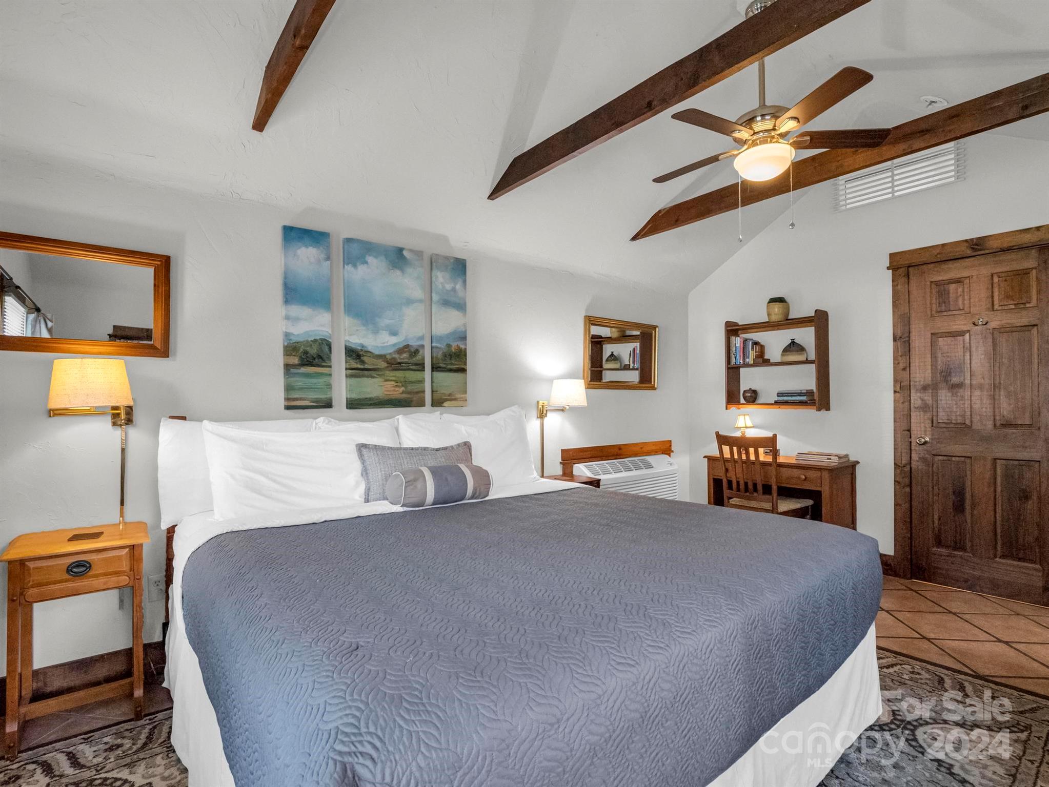 Furnished bedroom with a large bed beneath exposed wood beams and a ceiling fan.