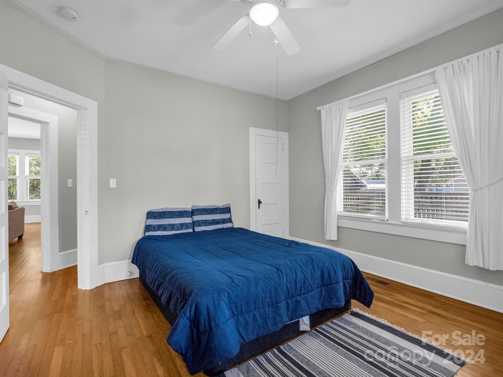 Furnished bedroom with a blue bed, hardwood floors, and two curtained windows.