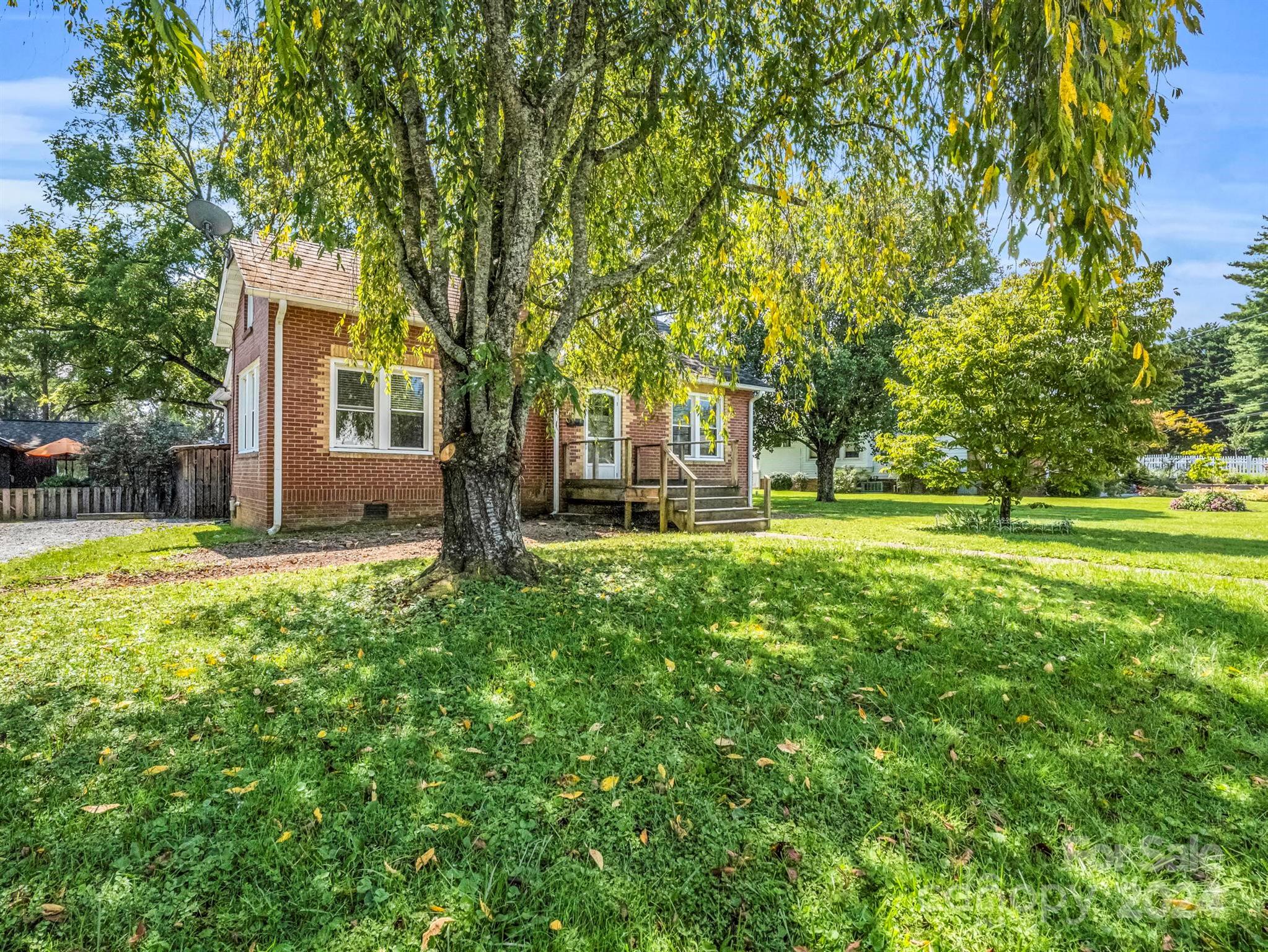 Red-brick house behind a large tree, with a wide lawn and a small wooden deck.